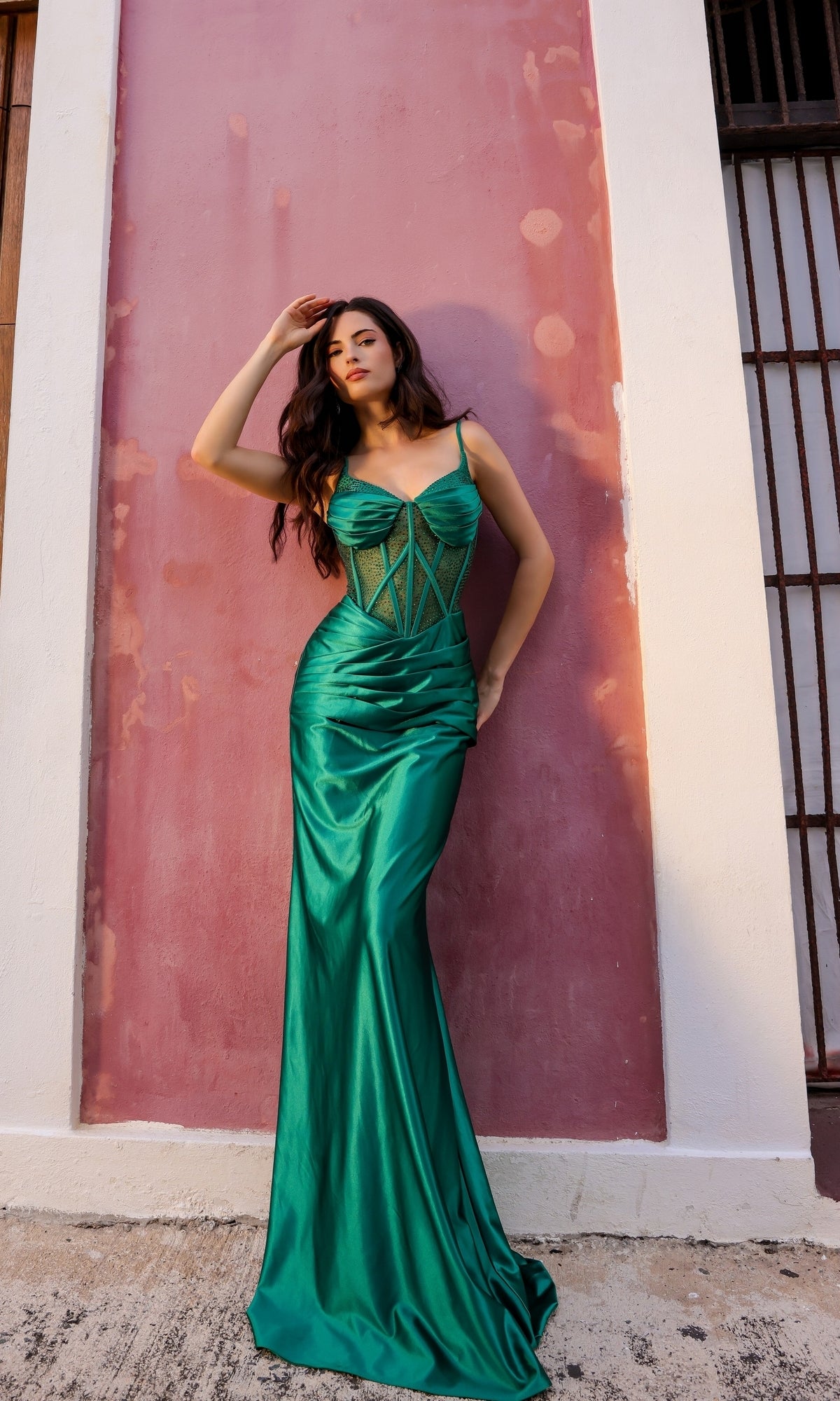 A woman poses against a pink and white wall, wearing the Lace-Up Corset-Bodice Long Satin Prom Dress E1292—a fitted, emerald green gown with a corset bodice—her hand gently touching her hair.