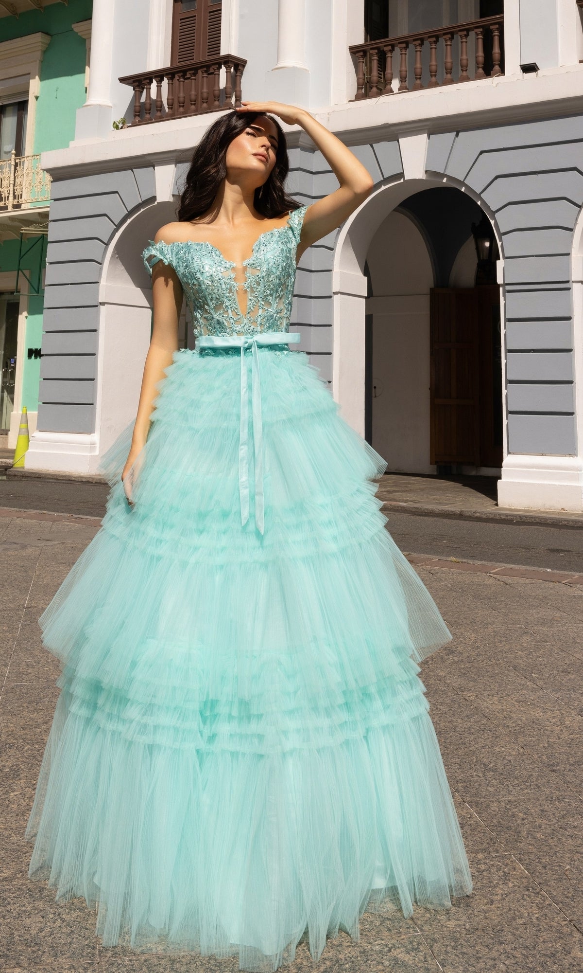 A woman stands outdoors, shielding her eyes from the sun, wearing the Nox Anabel Designer Ruffled Prom Ball Gown E1293 in light blue with beaded embroidered lace details and an off-the-shoulder design.