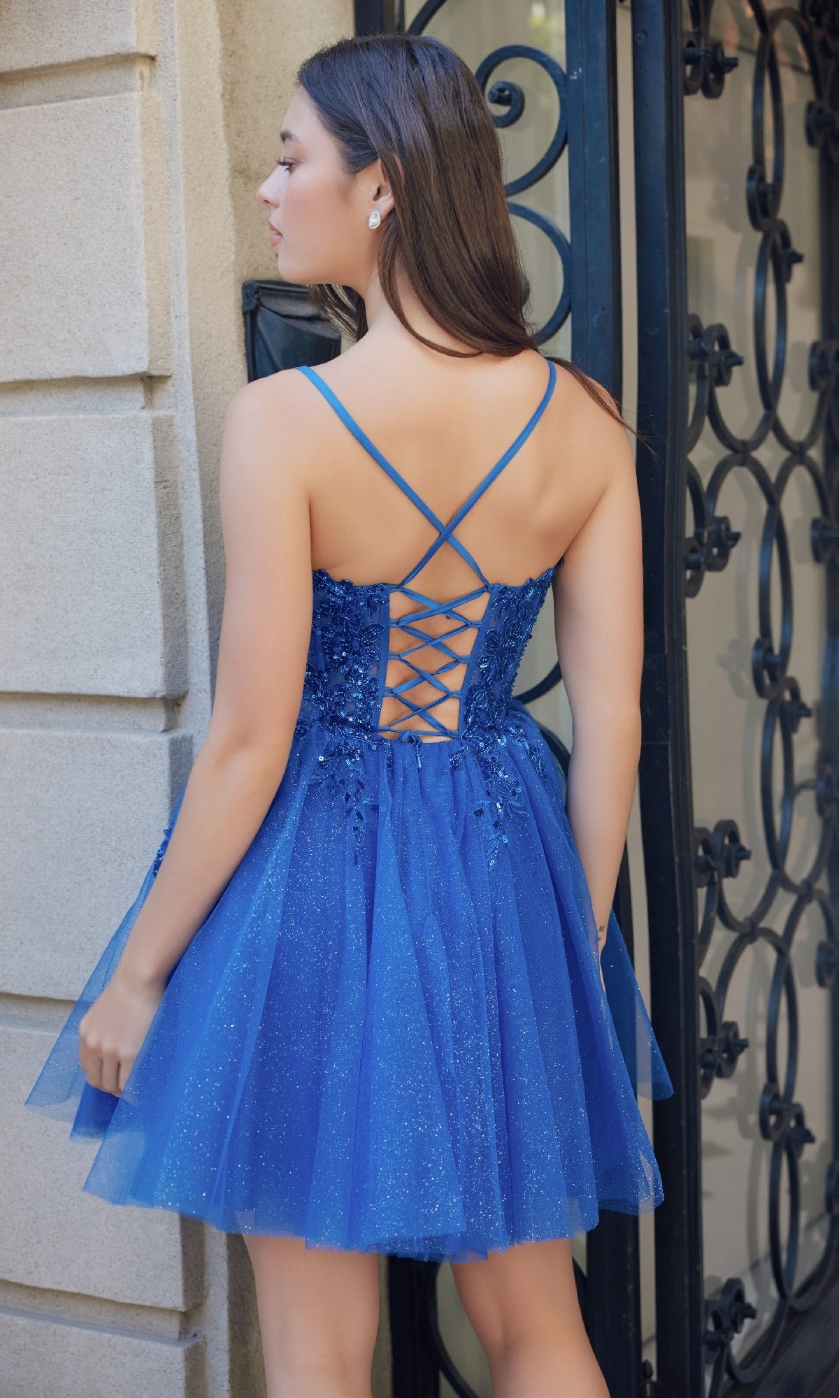 A woman in the Nox Anabel E902 Glitter Tulle Homecoming Dress, featuring sequin embroidery and a lace-up back, stands by a stone wall and black iron gate, facing away from the camera.