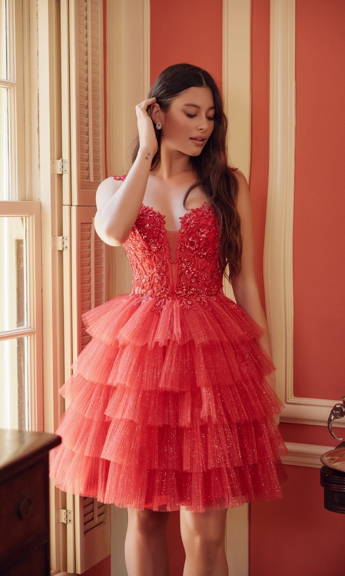 A woman in the Nox Anabel E904 Glitter Babydoll Homecoming Dress stands indoors by a window, touching her ear and looking down.