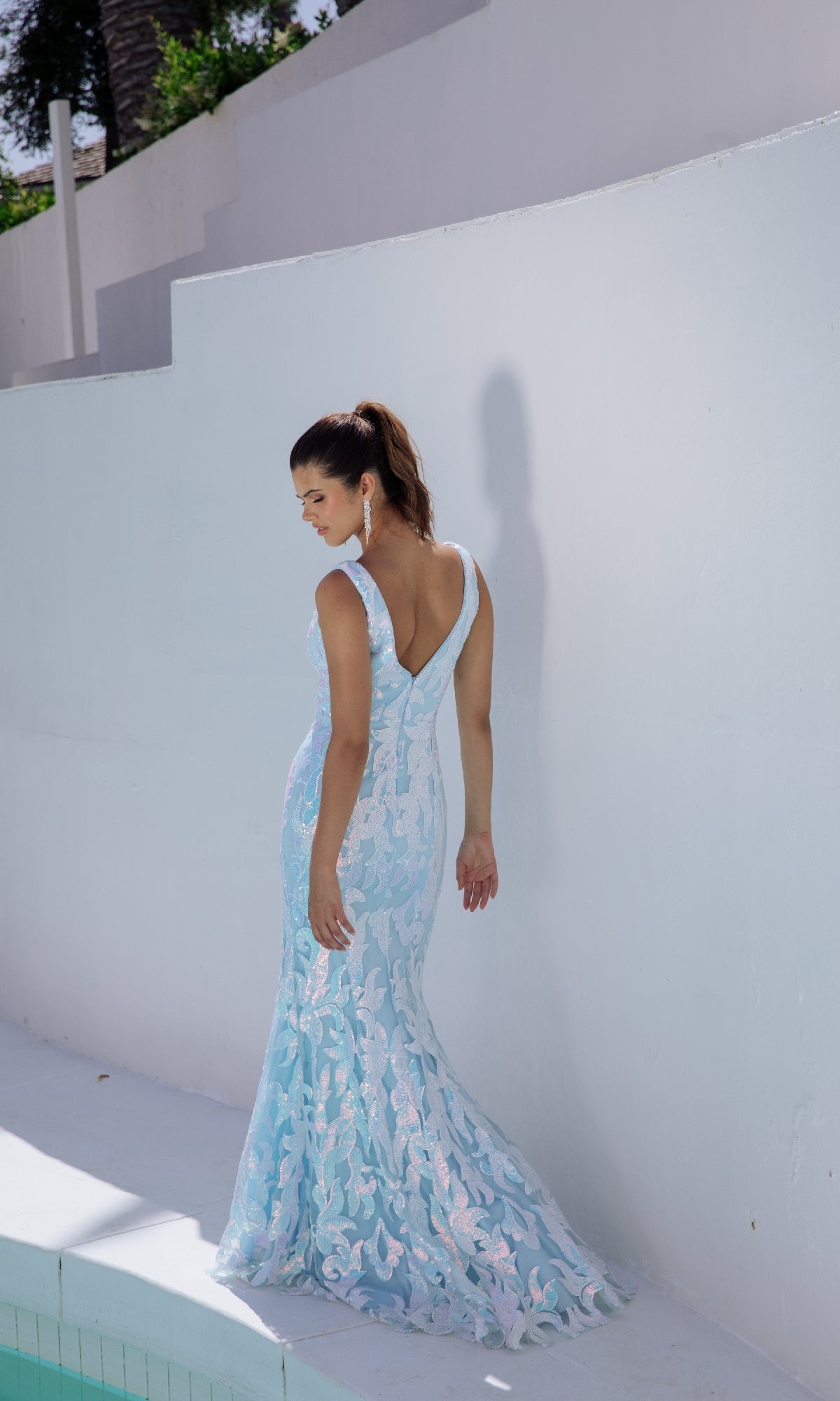 A woman in the Eureka EK101 V-Back Long Sequin-Print Prom Dress stands by a white curved wall, gazing down over her shoulder.