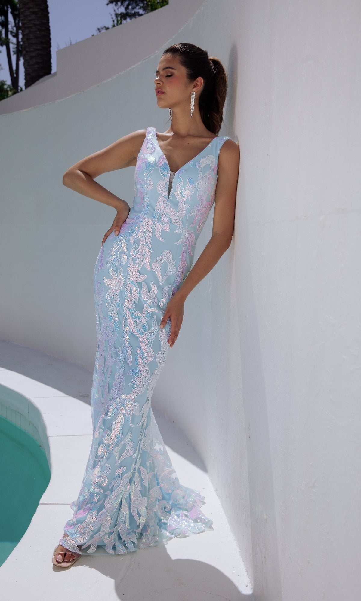 A woman in the V-Back Long Sequin-Print Prom Dress: Eureka EK101 stands by a white curved wall near a pool, looking to the side with one hand on her hip.