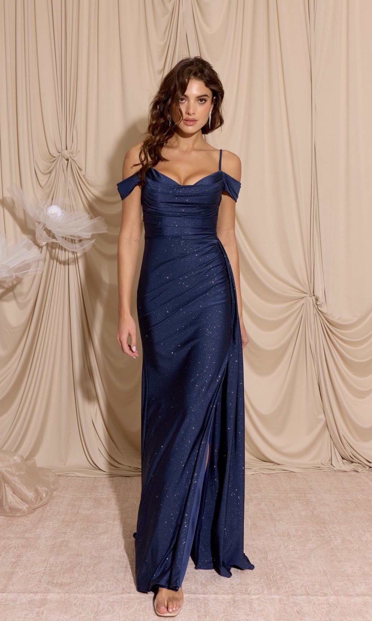 A woman poses before beige drapes in the Long Ruched Jersey Prom Dress: Eureka EK107—a fitted, navy blue, off-the-shoulder gown with a thigh-high slit that highlights her silhouette.