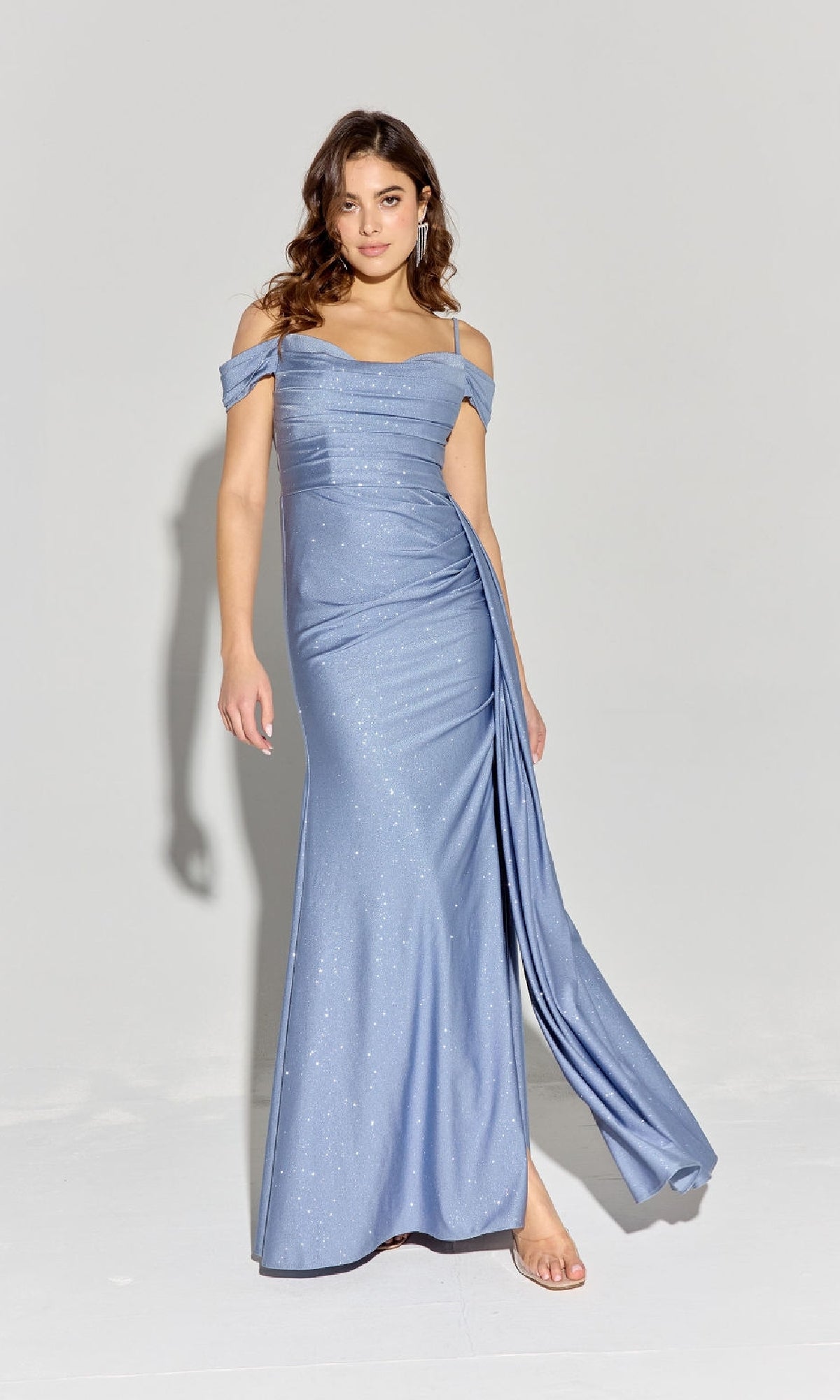 A woman wears the Long Ruched Jersey Prom Dress: Eureka EK107 in light blue, featuring a shimmery off-the-shoulder design with a side drape, standing against a plain light background.