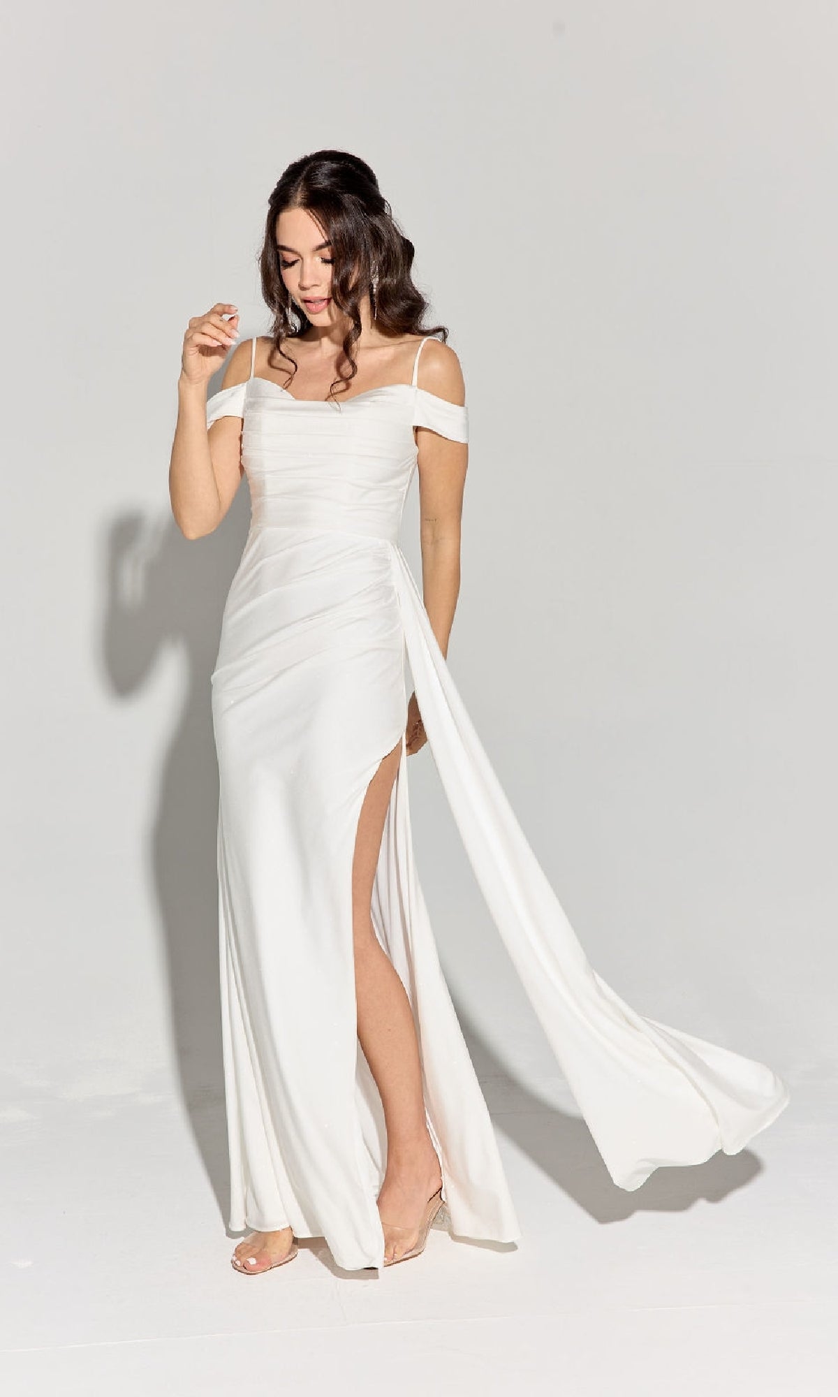 A woman models the Long Ruched Jersey Prom Dress: Eureka EK107, featuring an off-the-shoulder design, high slit, and draped train, against a plain light background.