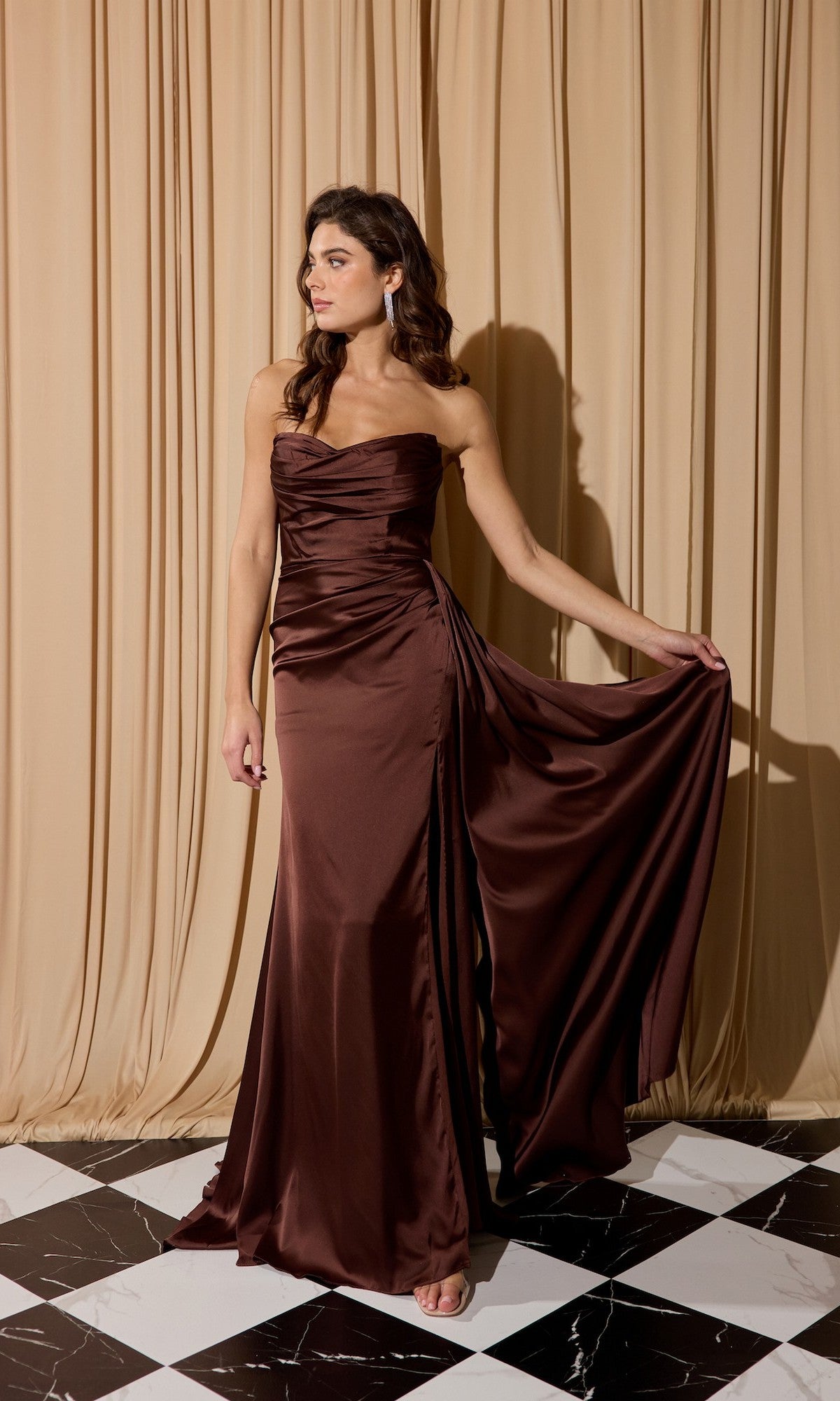 A woman stands on a black and white checkered floor, wearing the Strapless Long Satin Prom Dress: Eureka EK118 and holding one side of the brown gown. Beige curtains hang in the background.