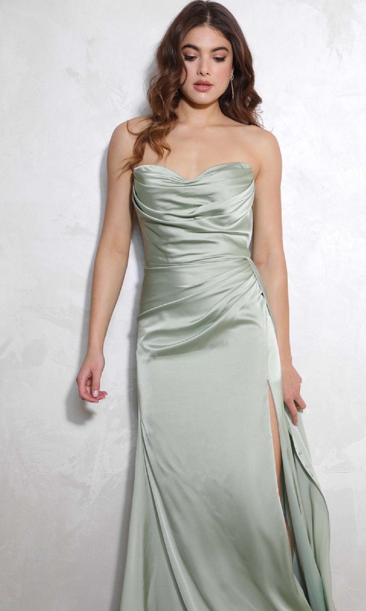 A woman in the Eureka EK118 Strapless Long Satin Prom Dress in light green, featuring a ruched bodice and thigh-high slit, stands against a light textured wall.