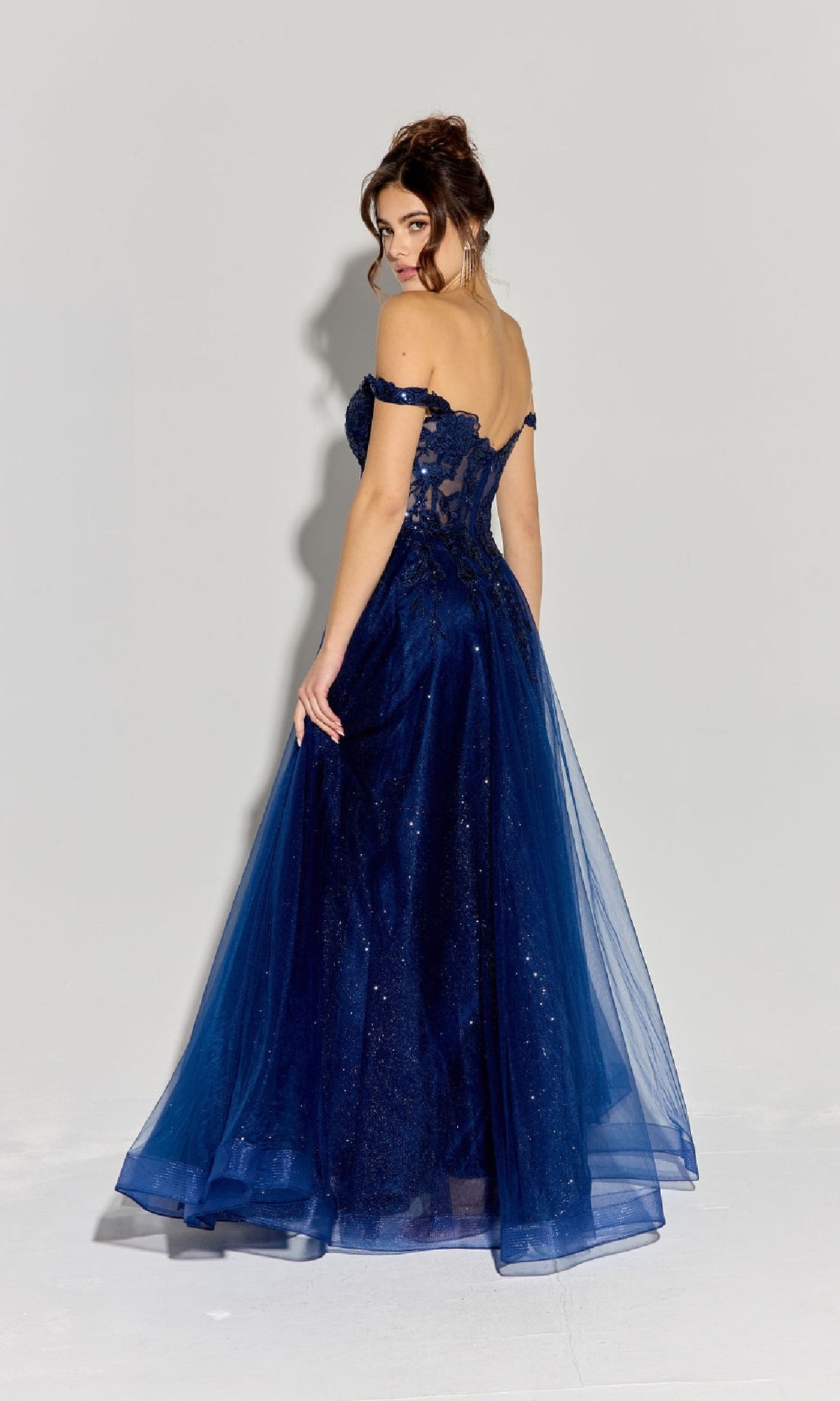 A woman in the Off-Shoulder Long Prom Ball Gown: Eureka EK128, featuring a sheer lace back and corset bodice in navy blue, stands facing away and glances over her shoulder against a plain background.
