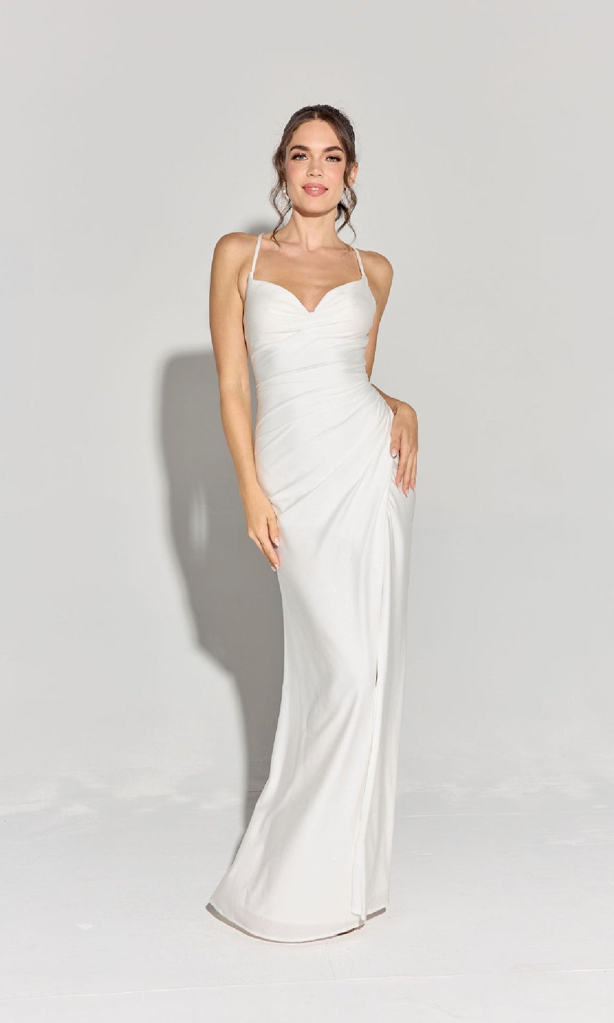 A woman stands against a plain background wearing the Classic Long Satin Prom Dress: Eureka EK136, a fitted white formal gown with thin straps and subtle waist draping—an elegant choice for any evening event.