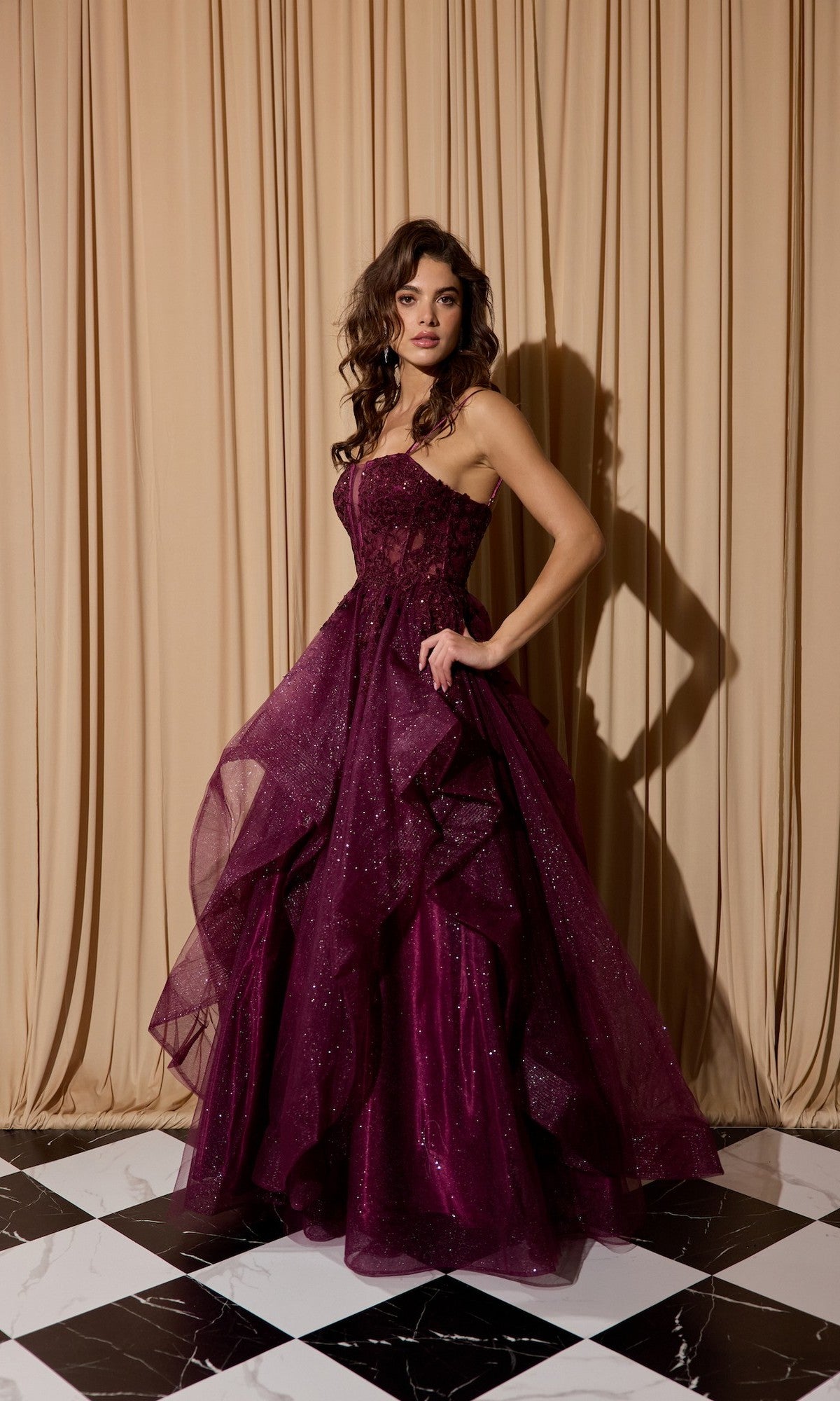 A woman in the Eureka EK157 long prom dress in berry, featuring a glitter tulle skirt, stands on a black and white checkered floor before beige curtains.