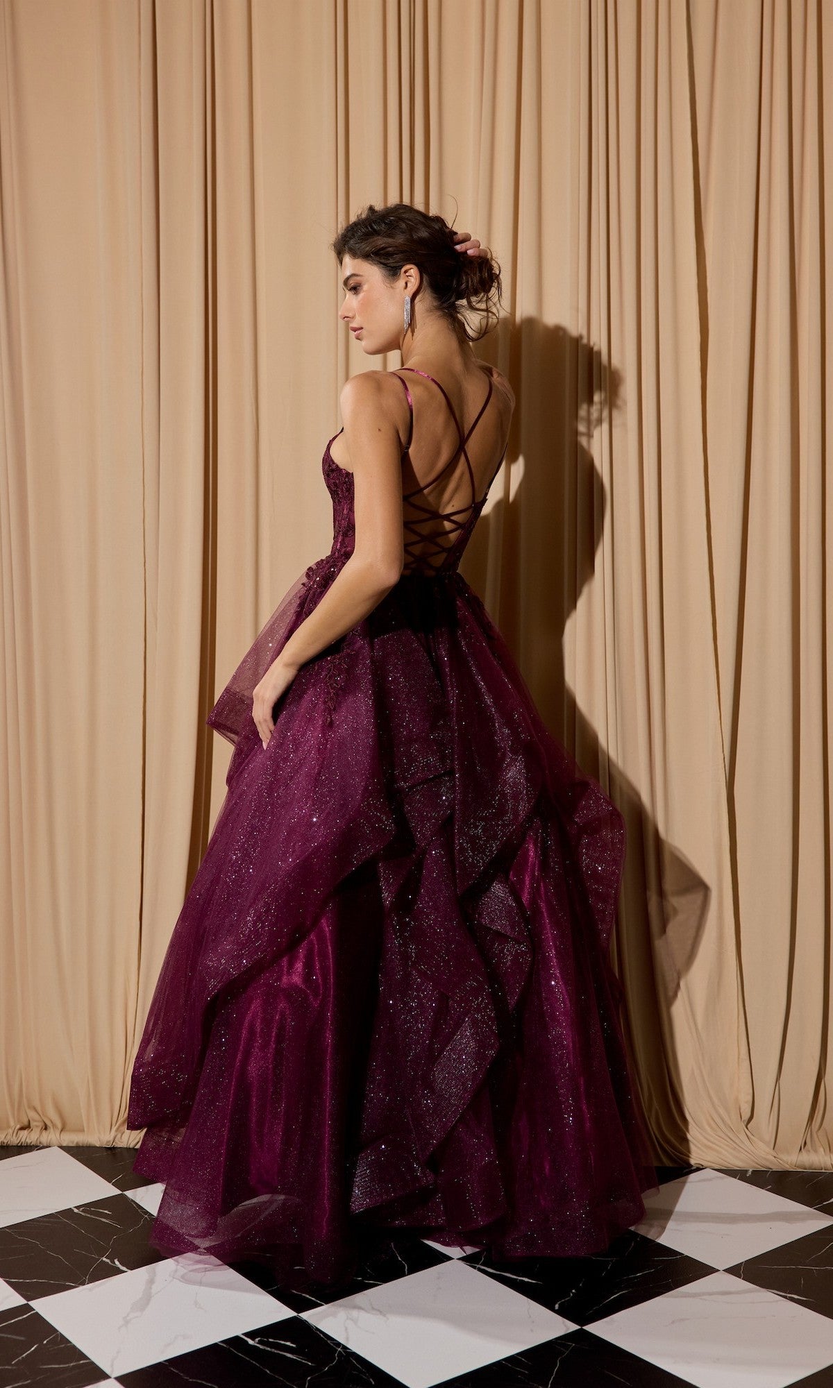 A woman wears the Eureka EK157 long prom dress in deep burgundy glitter, featuring a lace-up open back, as she stands on a black-and-white checkered floor before beige curtains.