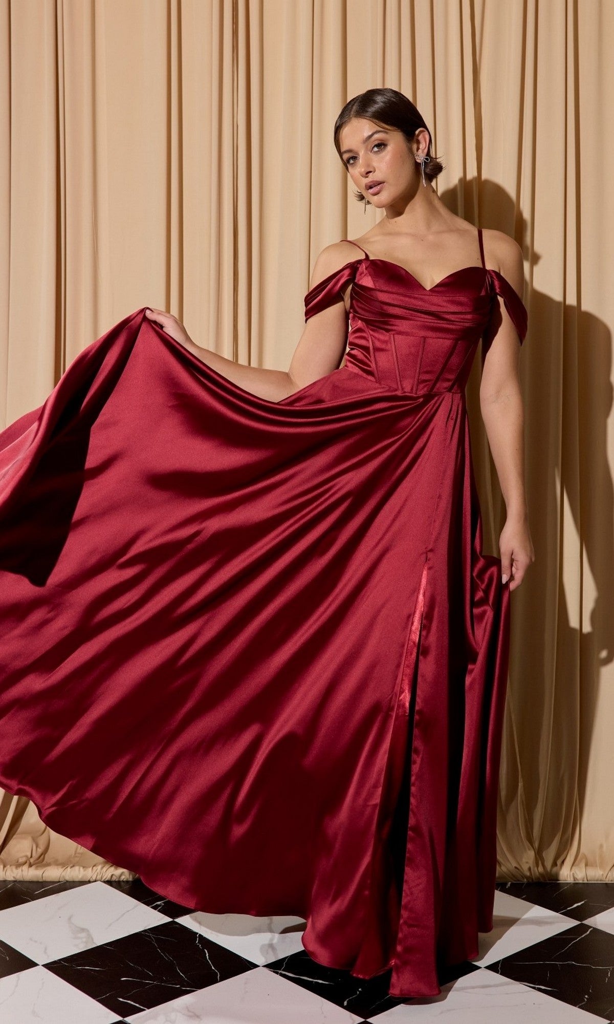A woman wears the Eureka EK176 long prom dress in burgundy, holding out the skirt while standing on a black and white tiled floor with beige curtains in the background.