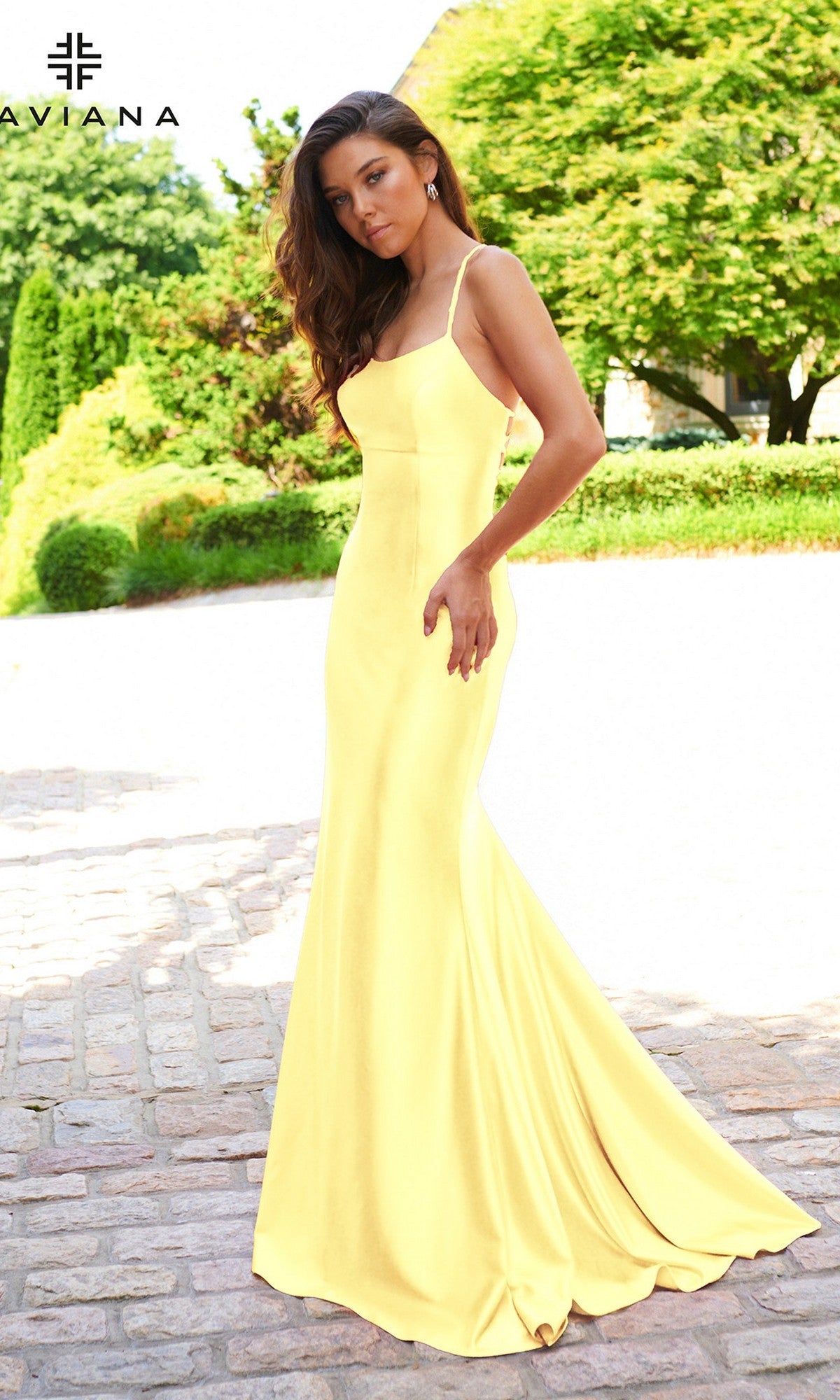 A woman stands outdoors in the Faviana ES10890, a Statement-Back long prom dress in yellow with thin straps, surrounded by lush green trees and a stone path.