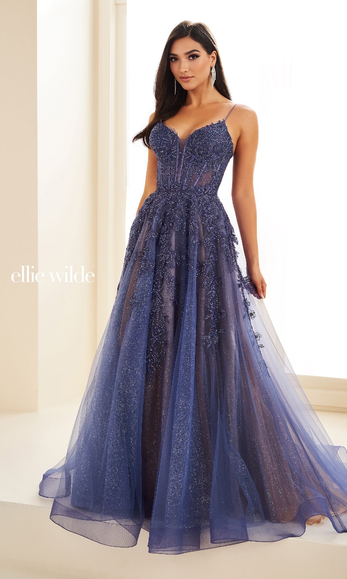 A woman stands indoors by a window wearing the Sheer-Corset Long Lace Prom Ball Gown EW34036, featuring thin straps, a fitted bodice, and an embellished tulle skirt.