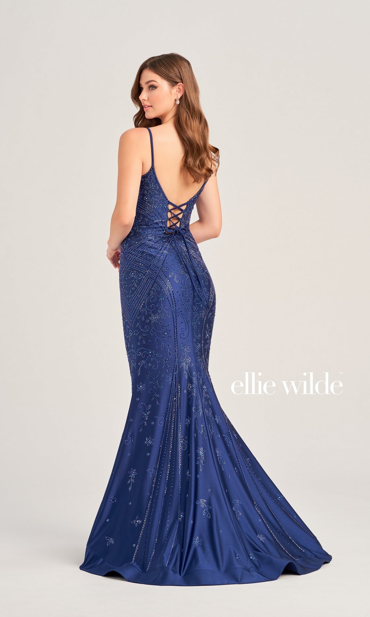 A woman wears the Ellie Wilde Long Designer Prom Dress EW35002—a fitted, floor-length deep blue mermaid gown with thin straps, a lace-up back, and sparkling beaded embellishments—standing and facing away.