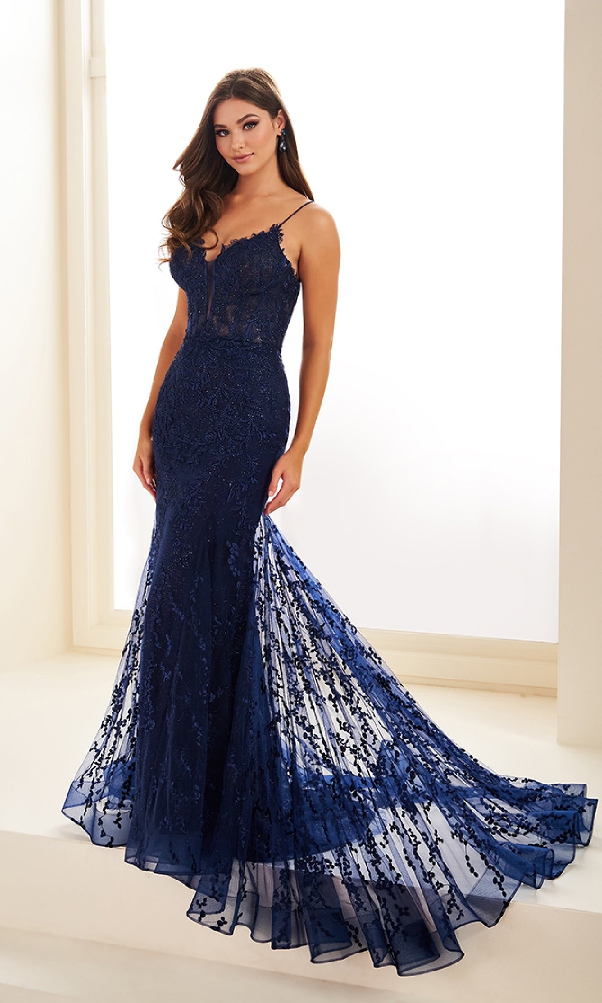 A woman wearing the Ellie Wilde Beaded-Tulle Long Prom Dress EW35010, a fitted navy blue gown with lace details and thin straps, stands indoors near a bright window.