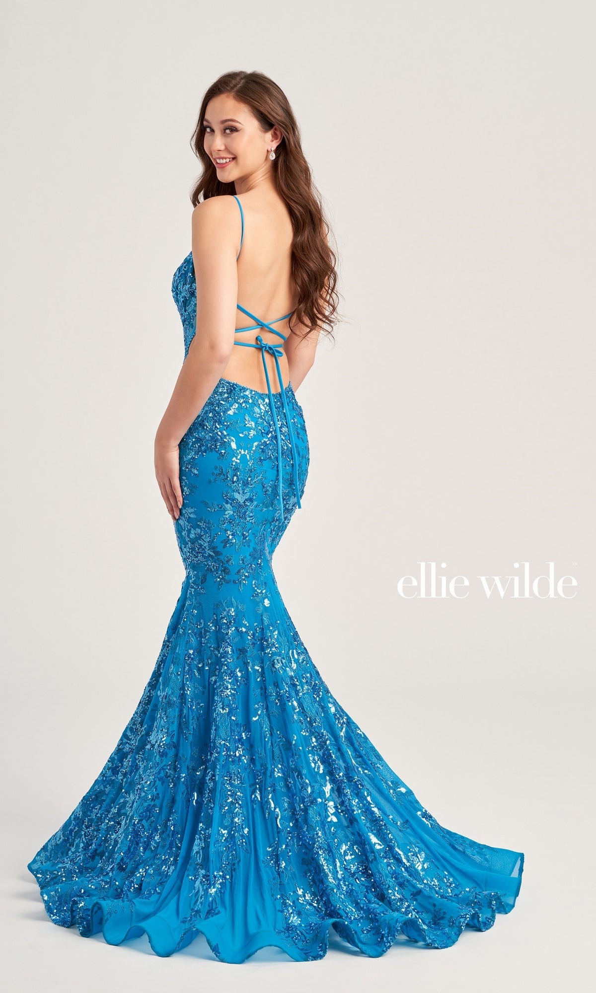 A woman in a bright blue sequin floral Ellie Wilde Long Tulle Designer Prom Dress EW35011 with an open back and crisscross straps stands facing away, turning slightly to smile over her shoulder. The