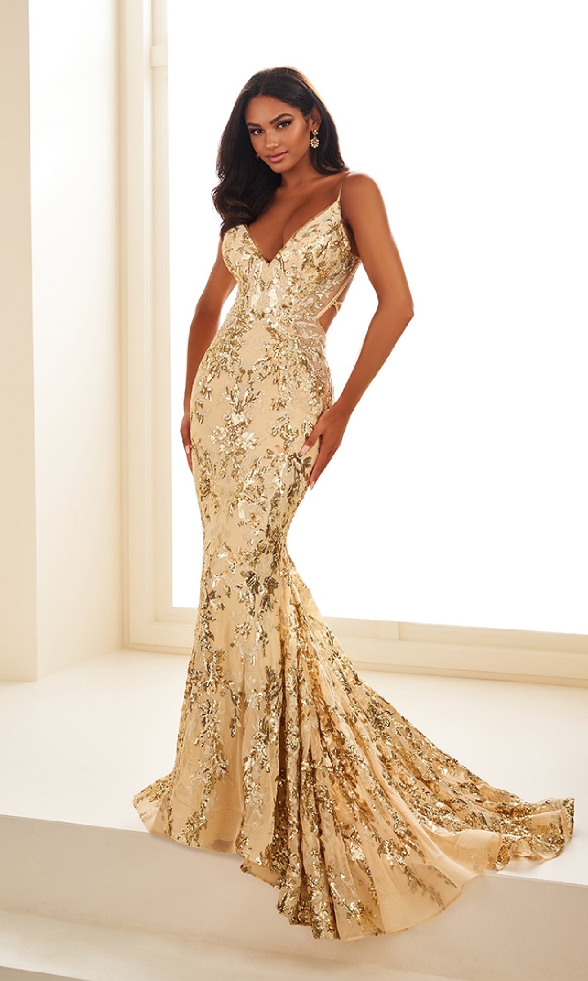 A woman stands indoors wearing the Ellie Wilde Long Tulle Designer Prom Dress EW35011—a glamorous, fitted, floor-length gold sequin floral gown with a train and deep V-neckline, perfect for prom night.