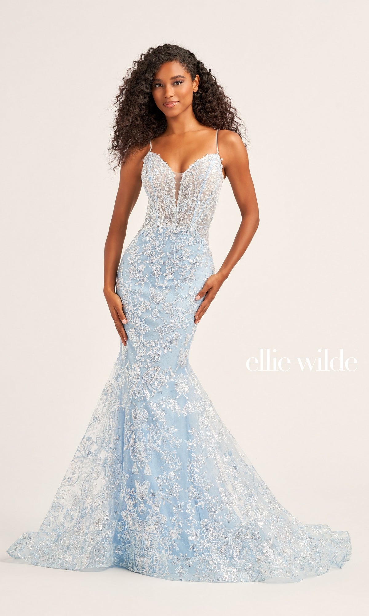 A woman poses in the Ellie Wilde Designer Glitter Prom Dress EW35013—a light blue, floor-length mermaid gown with silver floral embellishments and thin straps—against a neutral background. The text