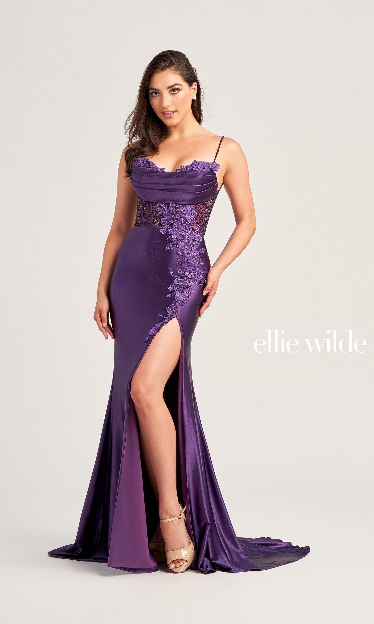 A woman in the Ellie Wilde Choker Designer Prom Dress EW35028—a floor-length purple gown with a thigh-high slit, beaded-lace floral appliques, and thin straps—stands against a plain background.