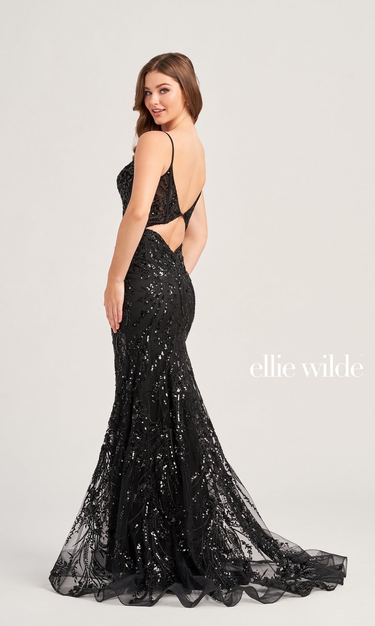 A woman models the Long Black Sequin Ellie Wilde Prom Dress EW35039, featuring thin straps, an open back, and a mermaid silhouette. She stands before a plain backdrop with