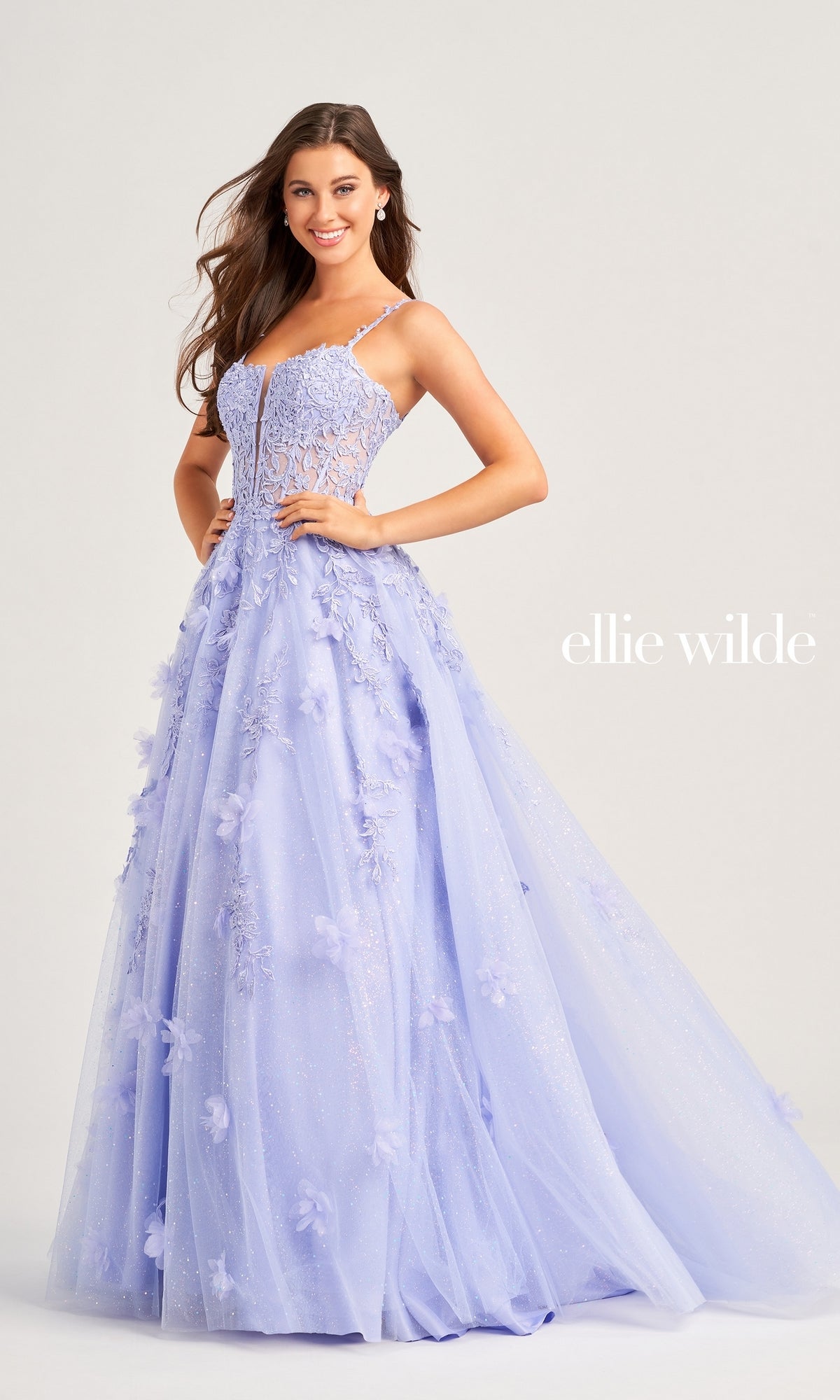 A woman in the Ellie Wilde Glitter-Tulle Long Prom Gown EW35081, featuring lavender floral appliqués and thin straps, stands smiling against a plain background.