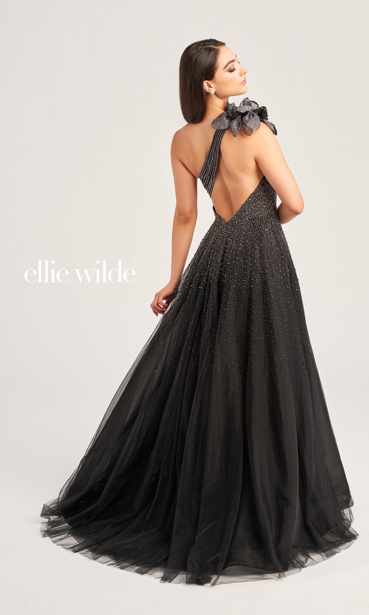 A woman models the Ellie Wilde Long Prom Dress EW35086, a black floor-length gown with an open back and single shoulder strap featuring a floral accent, standing against a plain background. The text