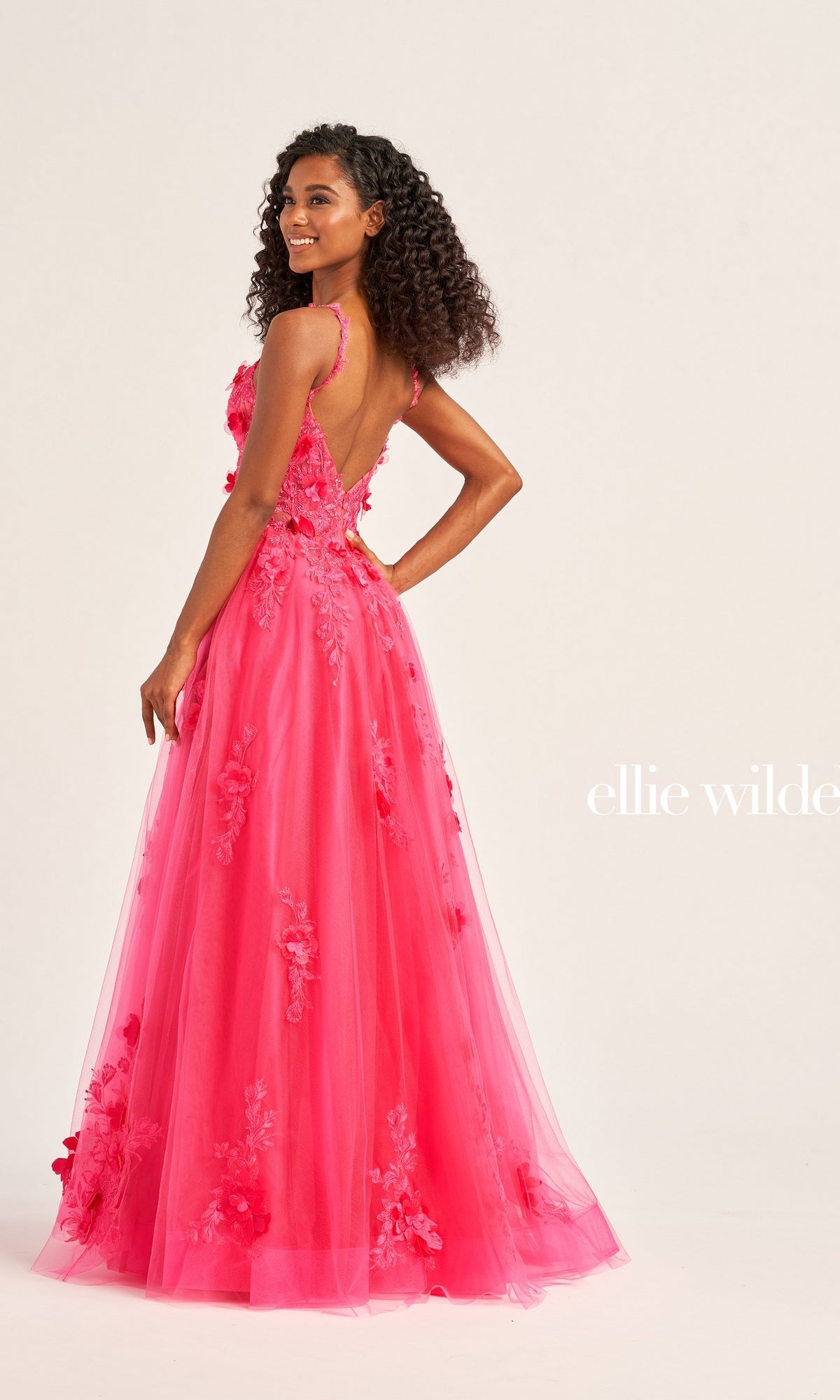 A woman smiles against a plain background wearing the Ellie Wilde Bright Pink Prom Ball Gown EW35122, featuring a floral-embellished corset bodice and low back.