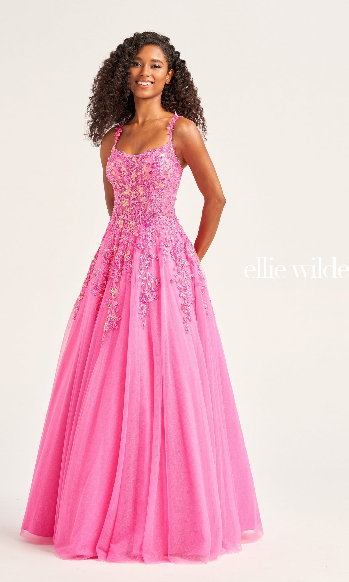 A woman wears the Ellie Wilde A-Line Long Designer Ball Gown EW35123, a bright pink dress with floral embellishments and thin straps, posed against a plain light background.