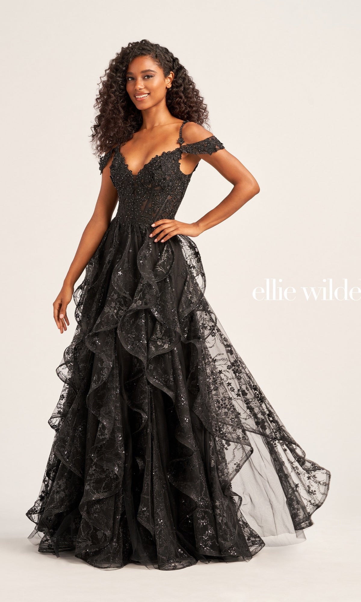 A woman wears the Ellie Wilde Ruffled Long Prom Ball Gown EW35218, a black, off-the-shoulder dress featuring ruffles, glittery details, and beaded lace appliques, while standing against a plain background.