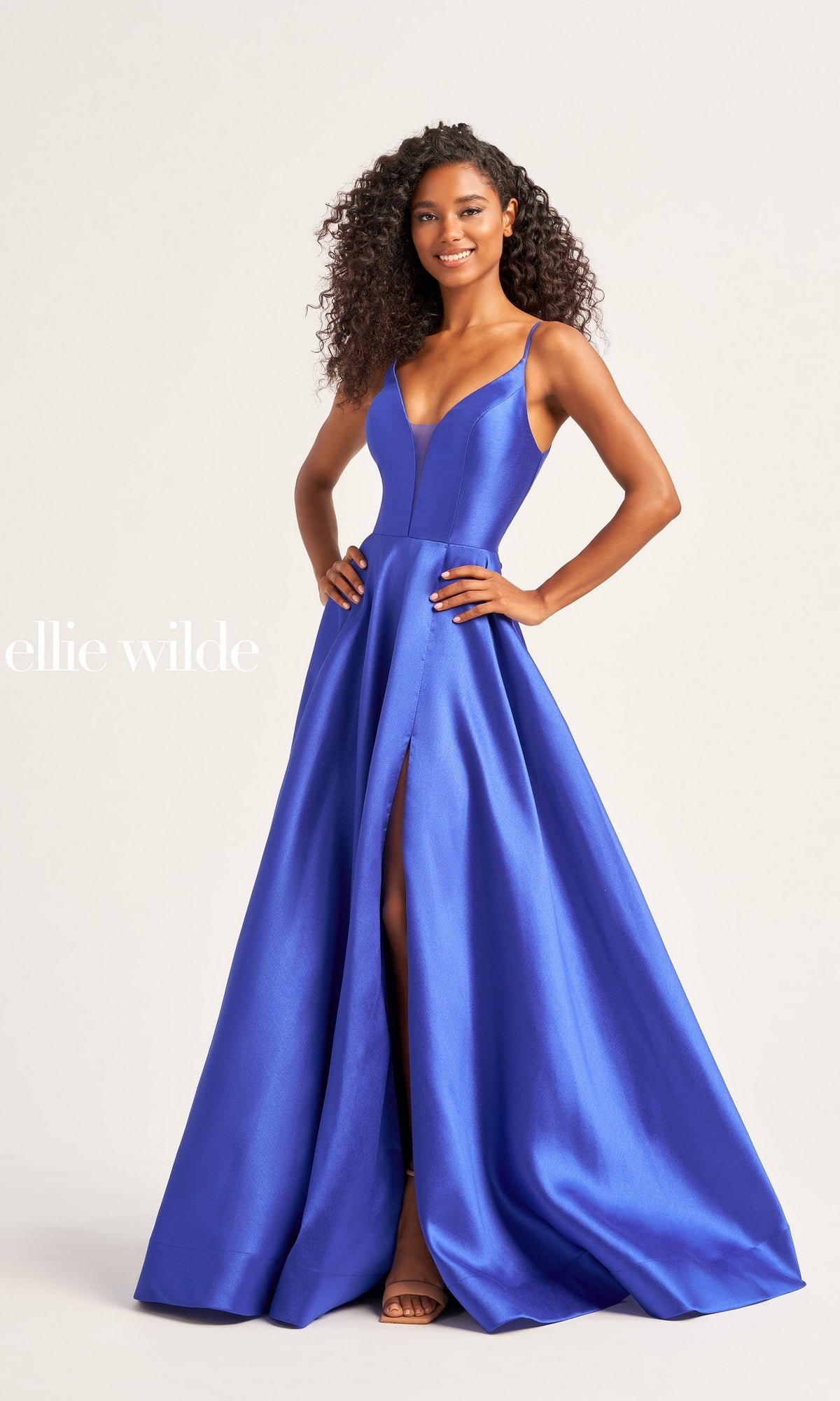 A woman smiles against a plain background in the Ellie Wilde Long A-Line Satin Prom Dress EW35232, a floor-length, sleeveless gown in deep blue with strappy back, thigh-high slit, fitted bodice, and flared skirt for an elegant evening look.
