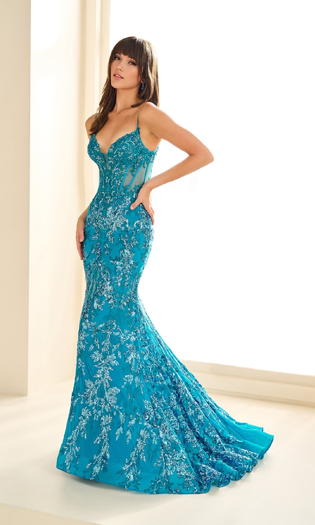 A woman in the Ellie Wilde EW36003 Sequin-Print Mermaid Prom Dress stands by a sunlit indoor window; the fitted, floor-length turquoise gown features intricate silver embroidery.