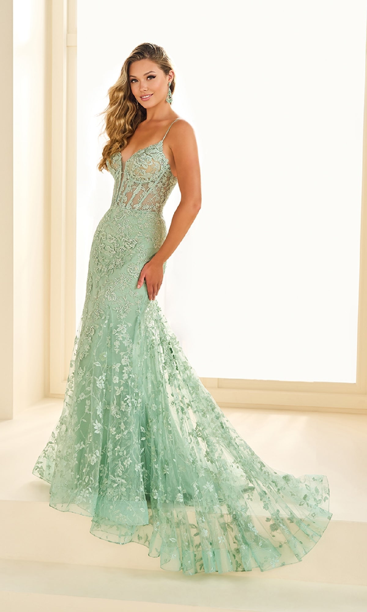 A woman wearing the Long Embroidered Prom Dress Ellie Wilde EW36008 in mint green, featuring a lace-up back, semi-sheer bodice, and flared floor-length skirt, standing indoors in natural light.
