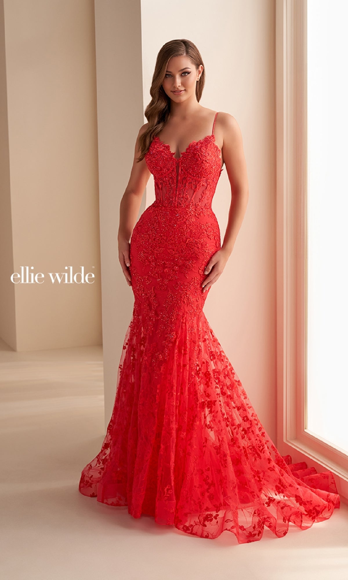 A woman stands in a softly lit room wearing the Ellie Wilde EW36008—a long red embroidered prom dress with thin straps, floral lace detailing, and a slight train. The