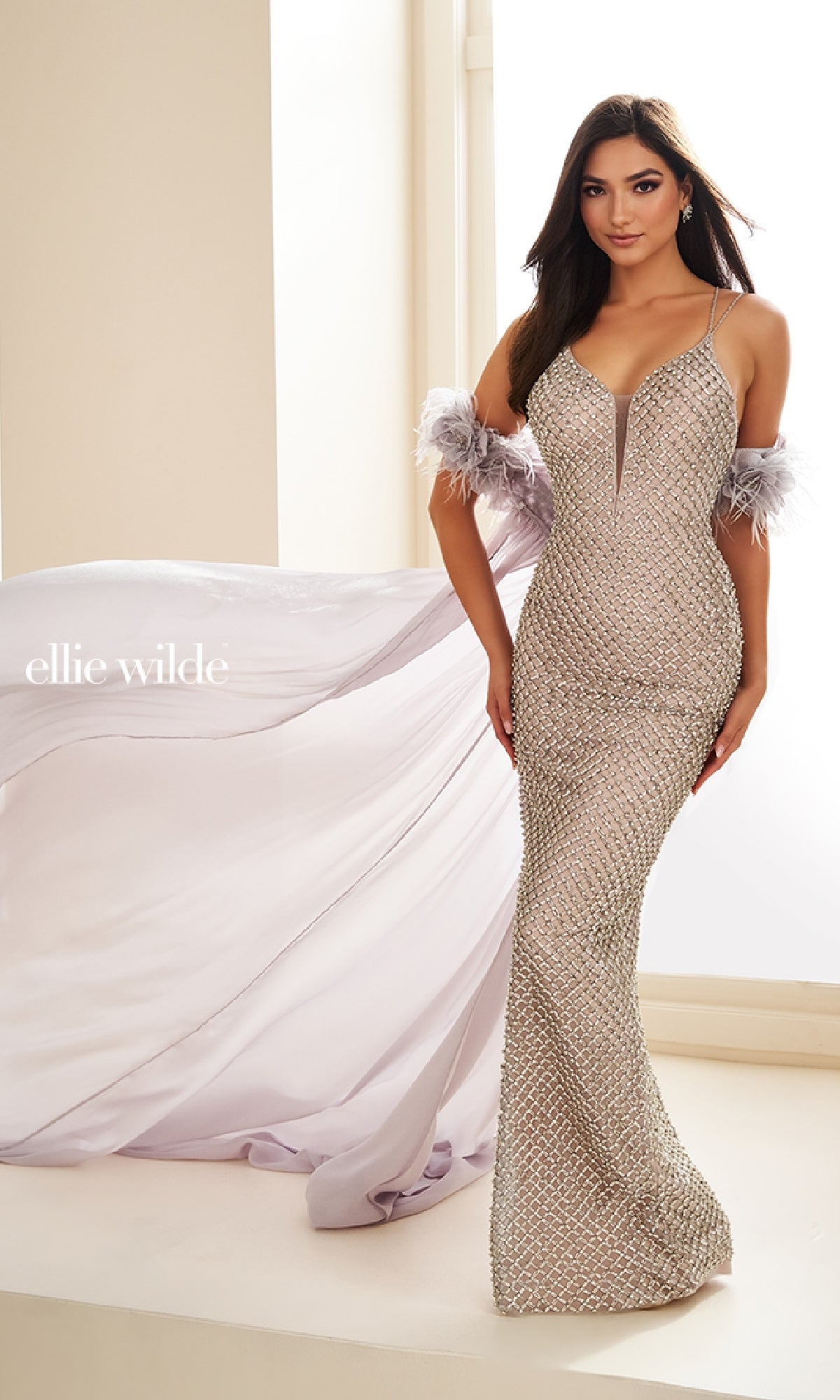 A woman wears the Ellie Wilde EW36027 long prom dress—fitted silver beaded mesh, feathery off-shoulder accents, and a detachable floor-length cape—while standing indoors.