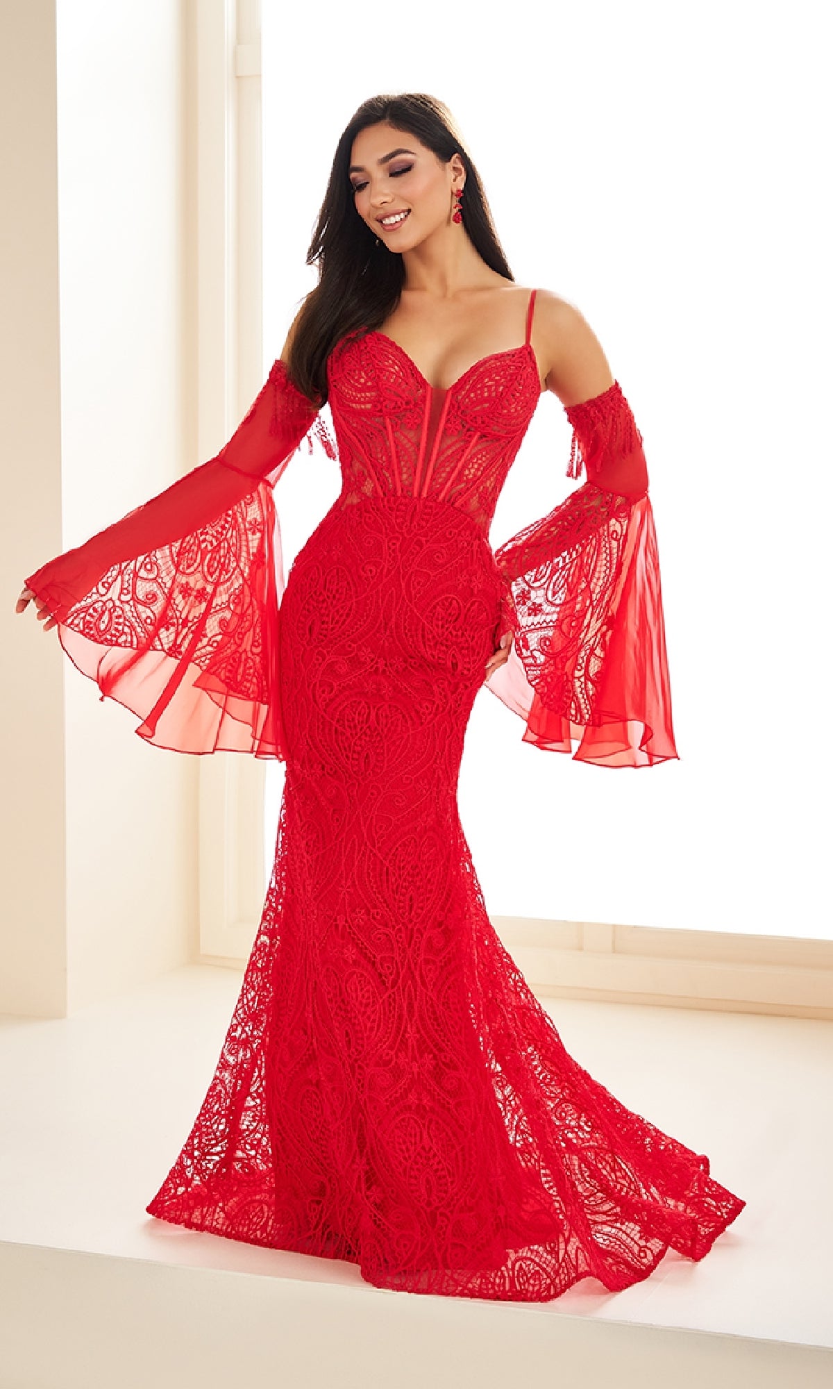 A woman stands indoors, smiling with arms raised, wearing the Ellie Wilde EW36030— a stunning long prom dress with Venice lace, a corset bodice, and a mermaid silhouette.