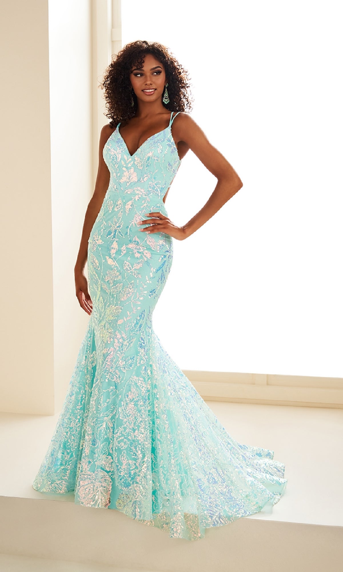 A woman poses indoors by a large window wearing the Ellie Wilde EW36034, a light blue, floor-length prom dress with floral sequins, thin straps, and a flared hem.