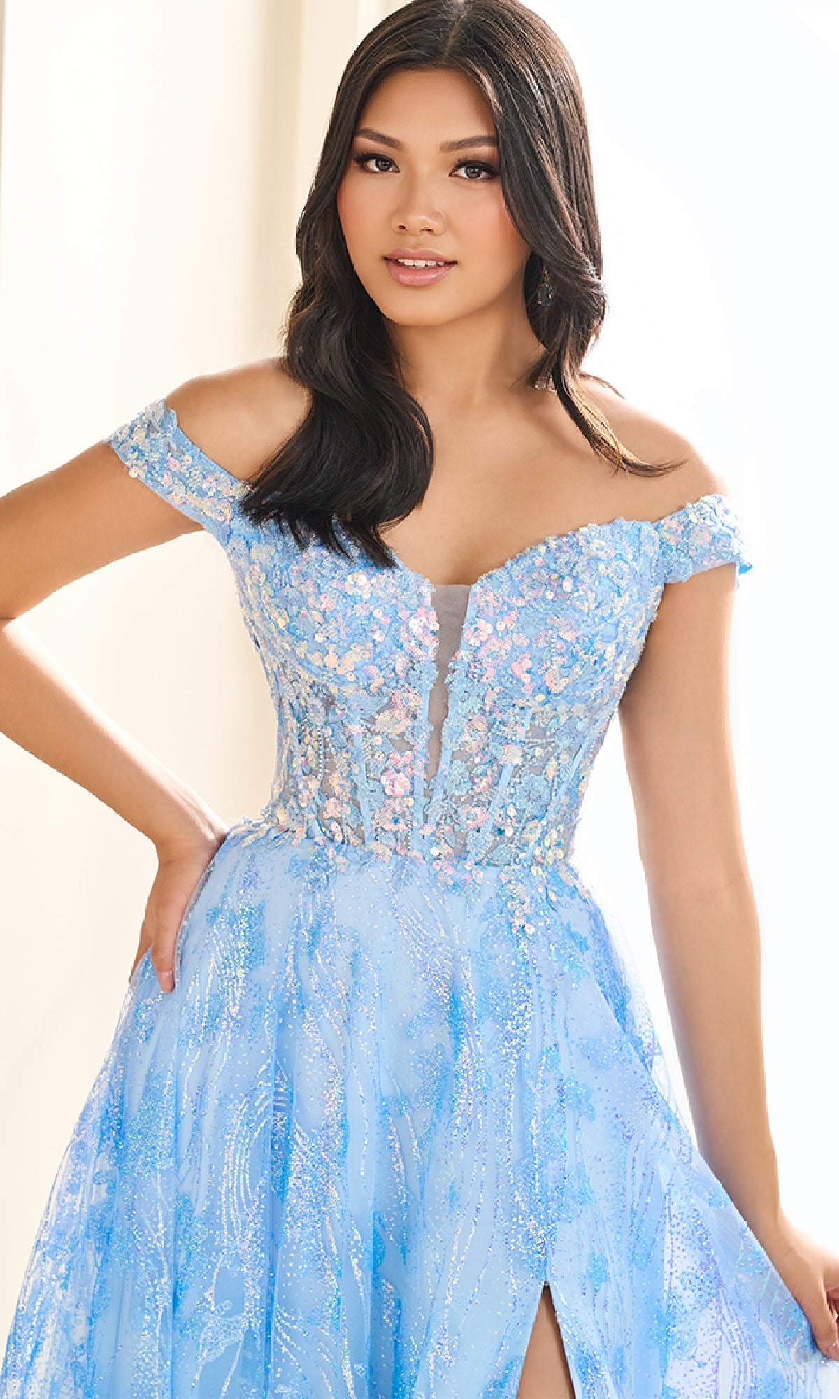 A woman stands indoors with one hand on her hip, wearing the Ellie Wilde EW36039 glow-in-the-dark prom dress featuring an off-the-shoulder design, light blue hue, floral embroidery, and a sheer corset bodice.