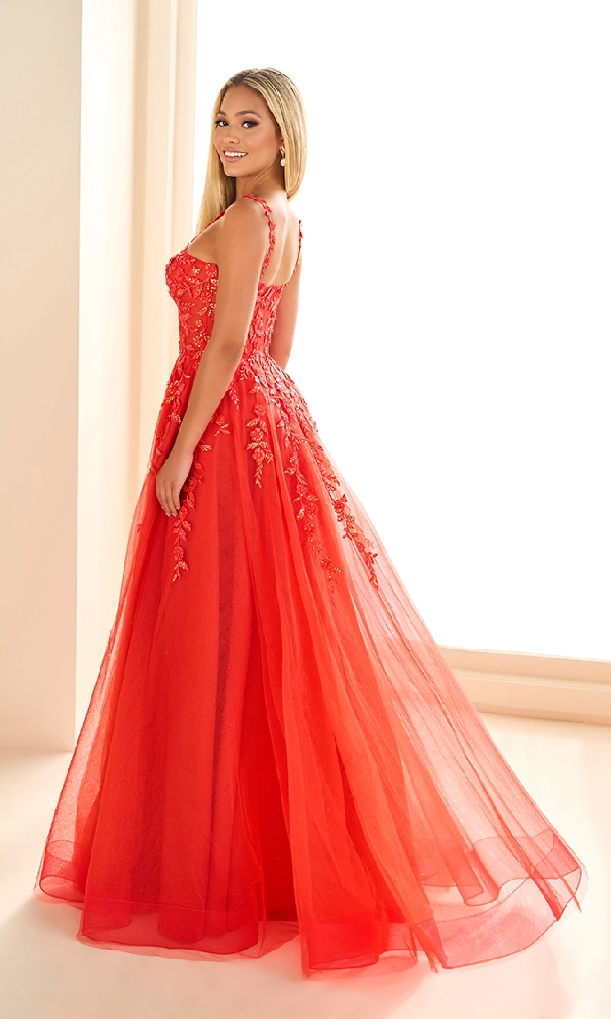 A woman in the Ellie Wilde EW36046 long prom dress, sleeveless and floor-length with floral embroidery, stands smiling and slightly turned to the side in a brightly lit room.