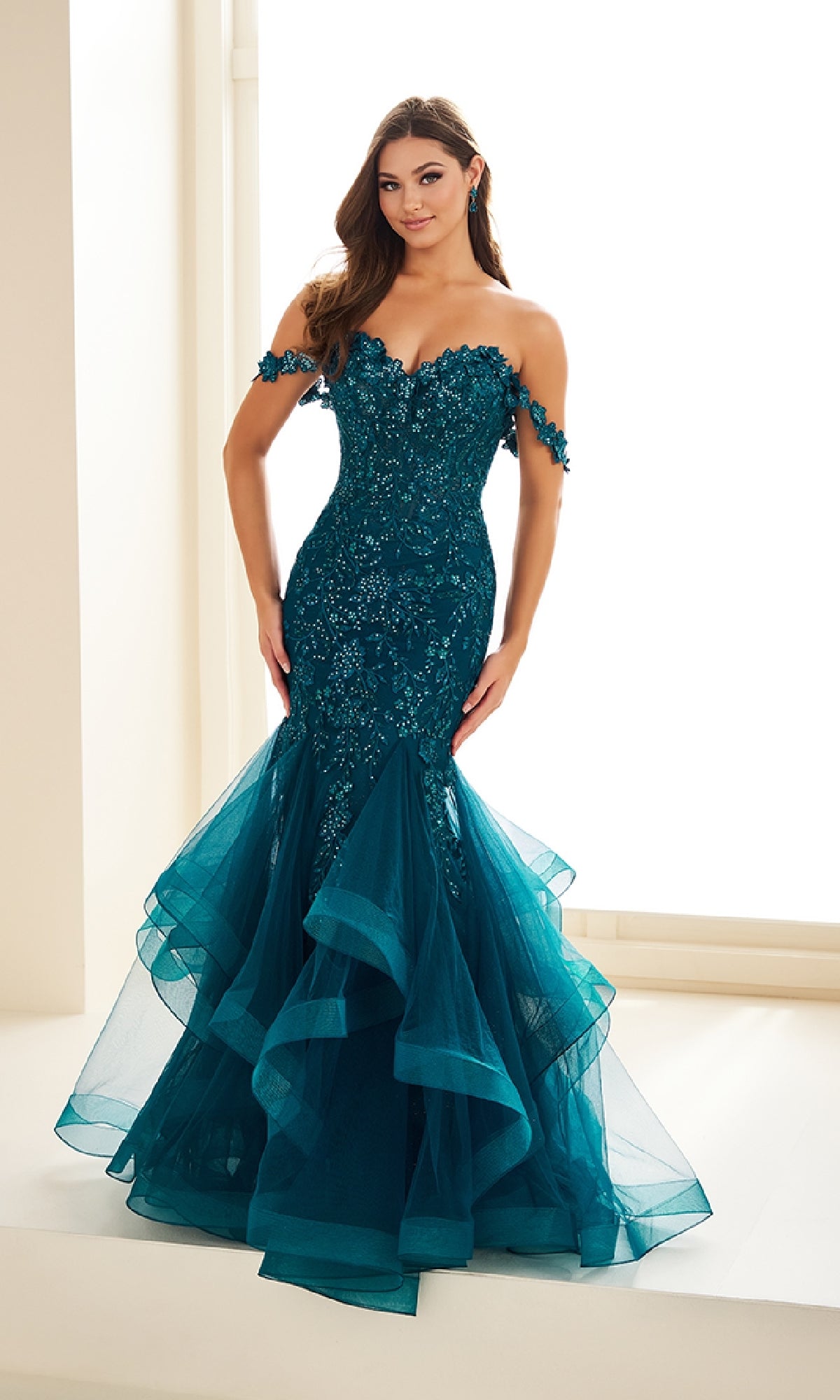 A woman stands indoors wearing the Ellie Wilde EW36047, a teal off-the-shoulder mermaid prom dress with sequins, glitter tulle ruffles, and floral appliqué detailing.