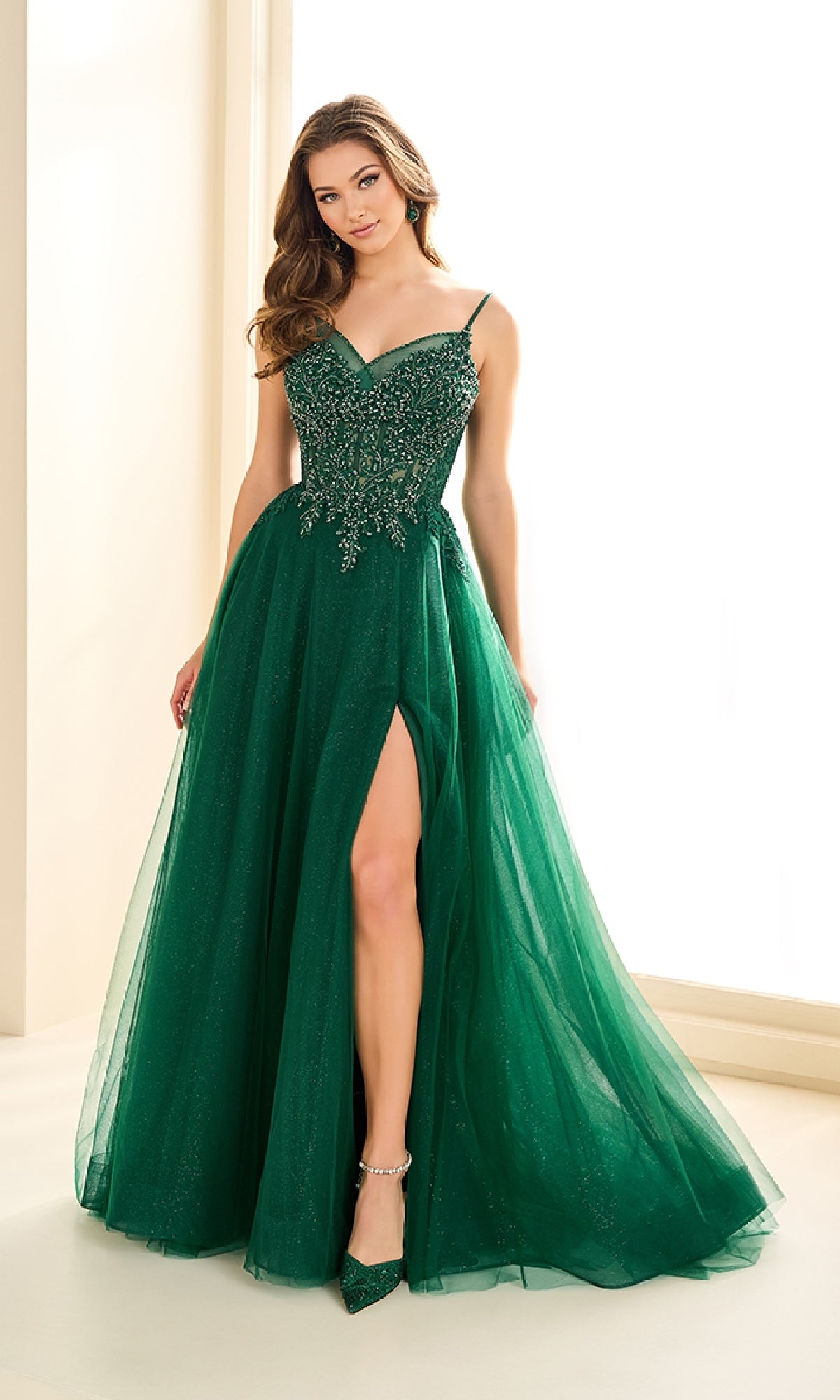 A woman poses indoors in the Ellie Wilde EW36066, a sleeveless dark green ball gown with an embellished corset bodice and high slit, holding her skirt with one hand.