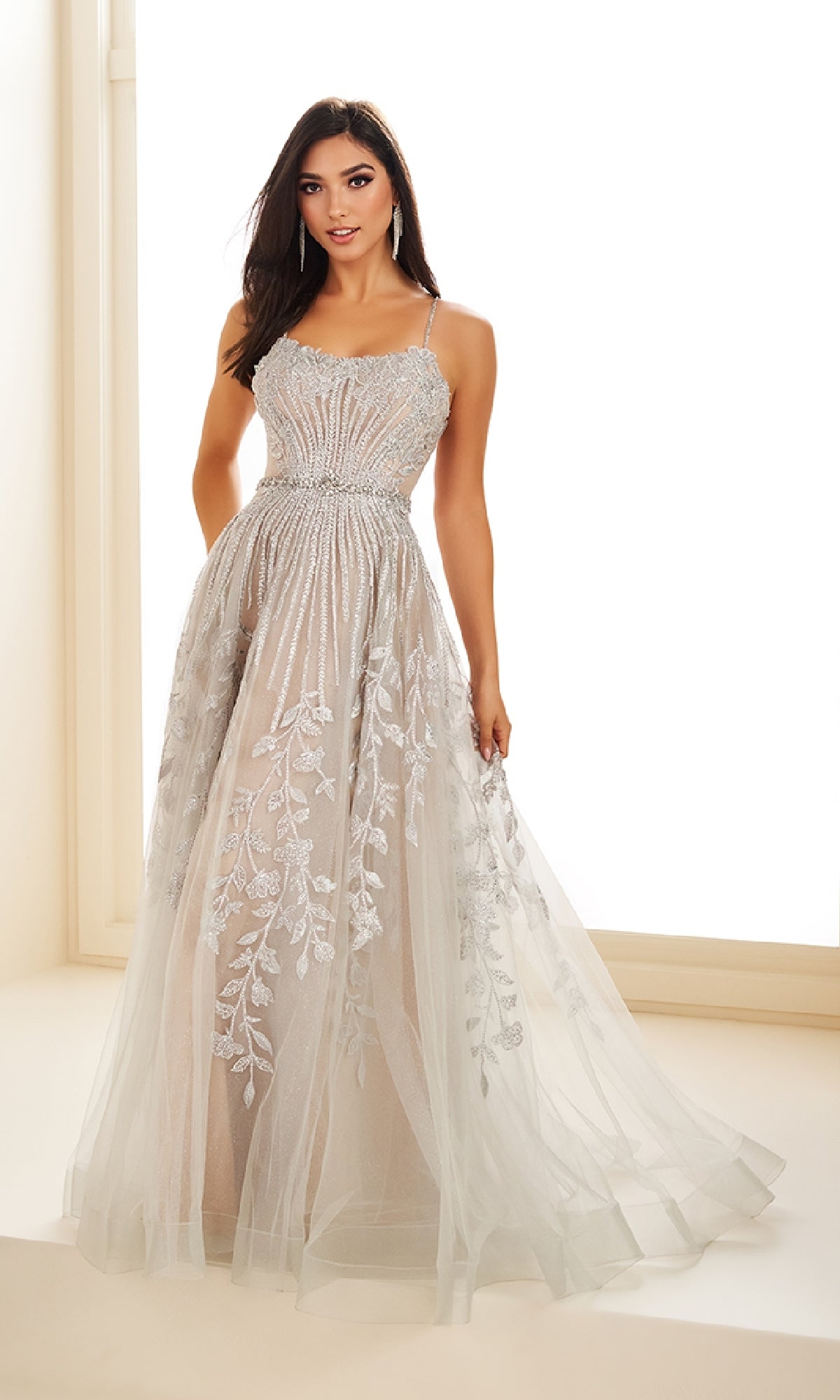 A woman poses in front of a bright window wearing the Ellie Wilde EW36068 long prom dress, featuring intricate beadwork and floral embroidery in shimmering silver.