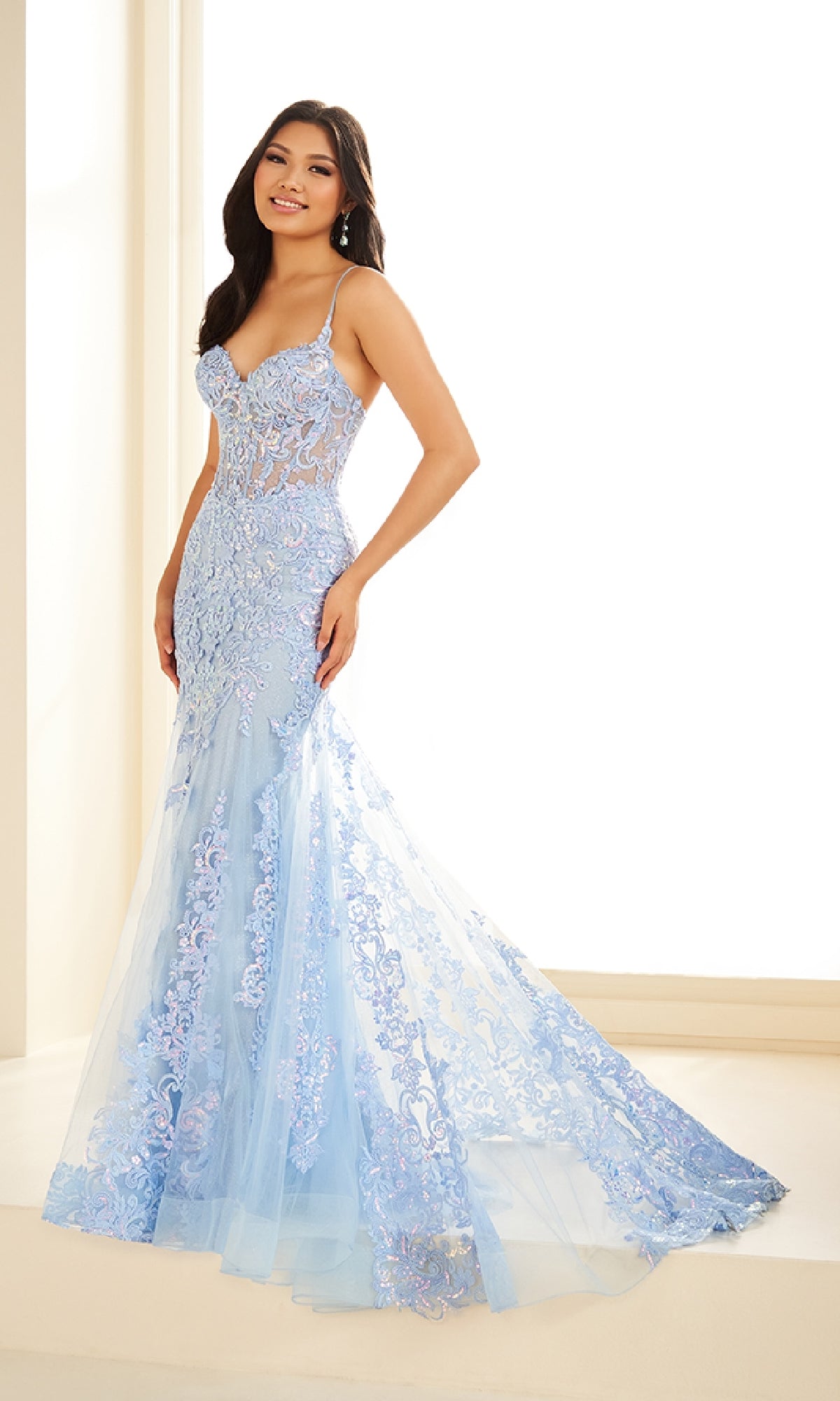 A woman poses indoors before a bright background, wearing the Ellie Wilde EW36069, a light blue, floor-length fitted prom dress with lace applique, thin straps, and glitter tulle in a glamorous mermaid style.