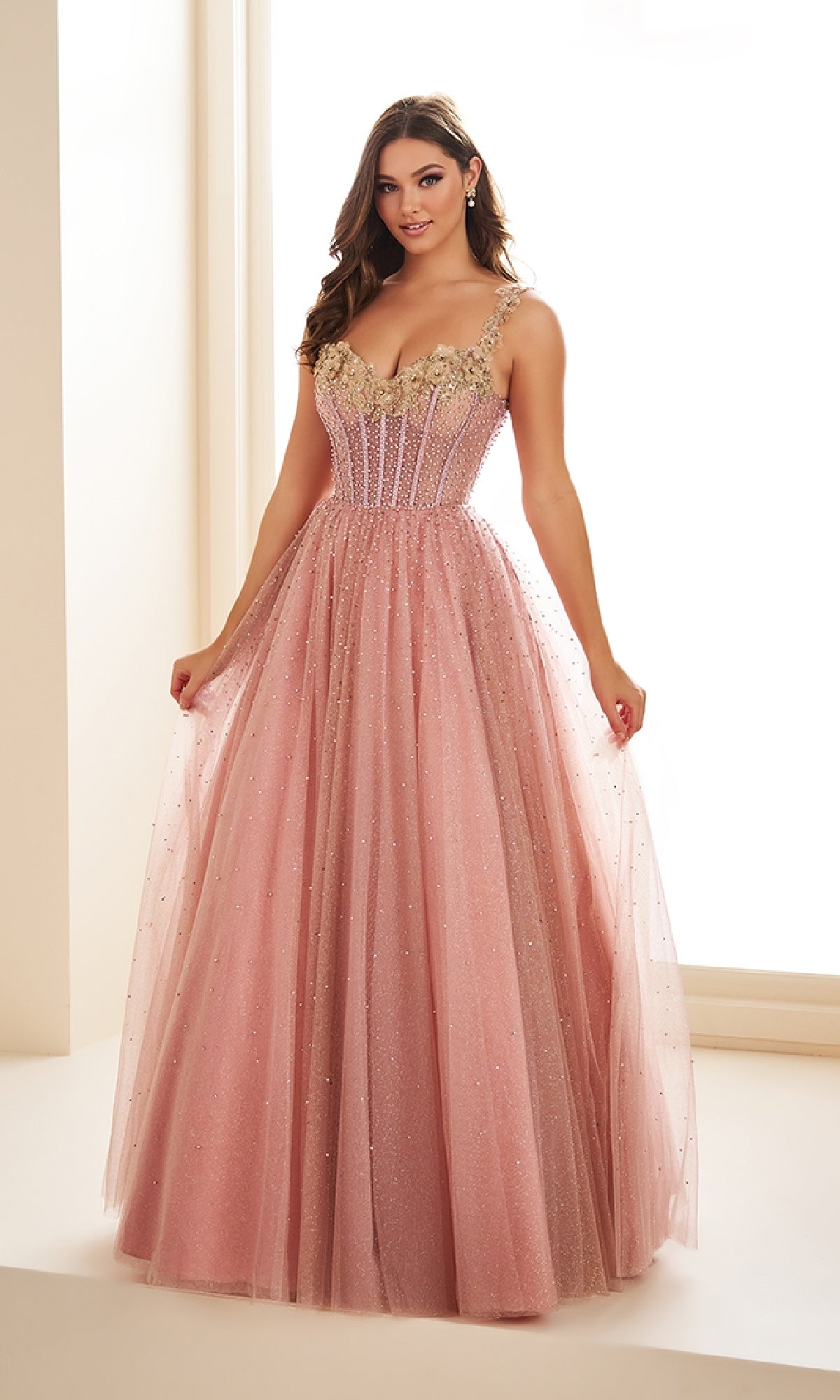 A woman stands indoors wearing the Long Prom Dress Ellie Wilde EW36071, a floor-length pink off-the-shoulder ball gown with floral appliqué straps, a fitted bodice, and a full glitter tulle skirt with sparkling embellishments.