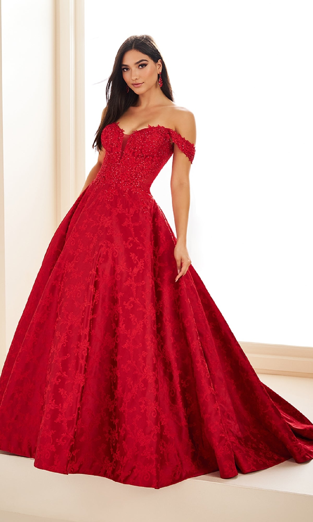 A woman poses in the Ellie Wilde EW36078, a floor-length, off-the-shoulder red prom dress featuring an embellished corset bodice and lace detailing, set in a bright, neutral space.