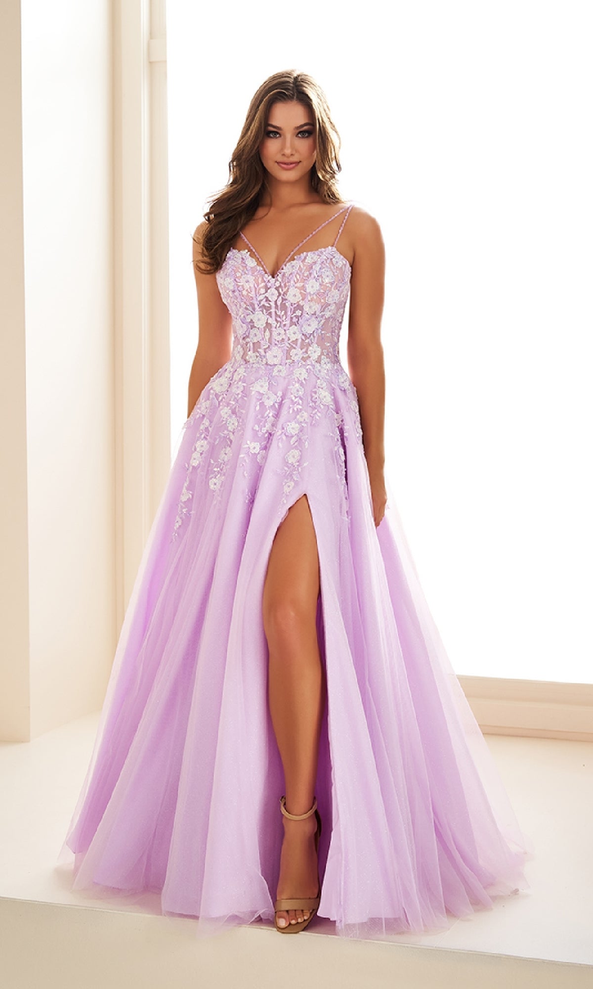 A woman stands indoors by a bright window wearing the Long Prom Dress: Ellie Wilde EW36079—a light purple gown with floral appliqué, thin straps, corset bodice, and a high slit.
