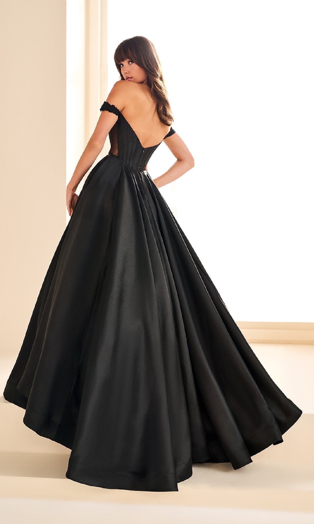 A woman stands near a bright window, looking over her shoulder while wearing the Ellie Wilde EW36100—a floor-length, off-the-shoulder prom ball gown with a fitted corset bodice.