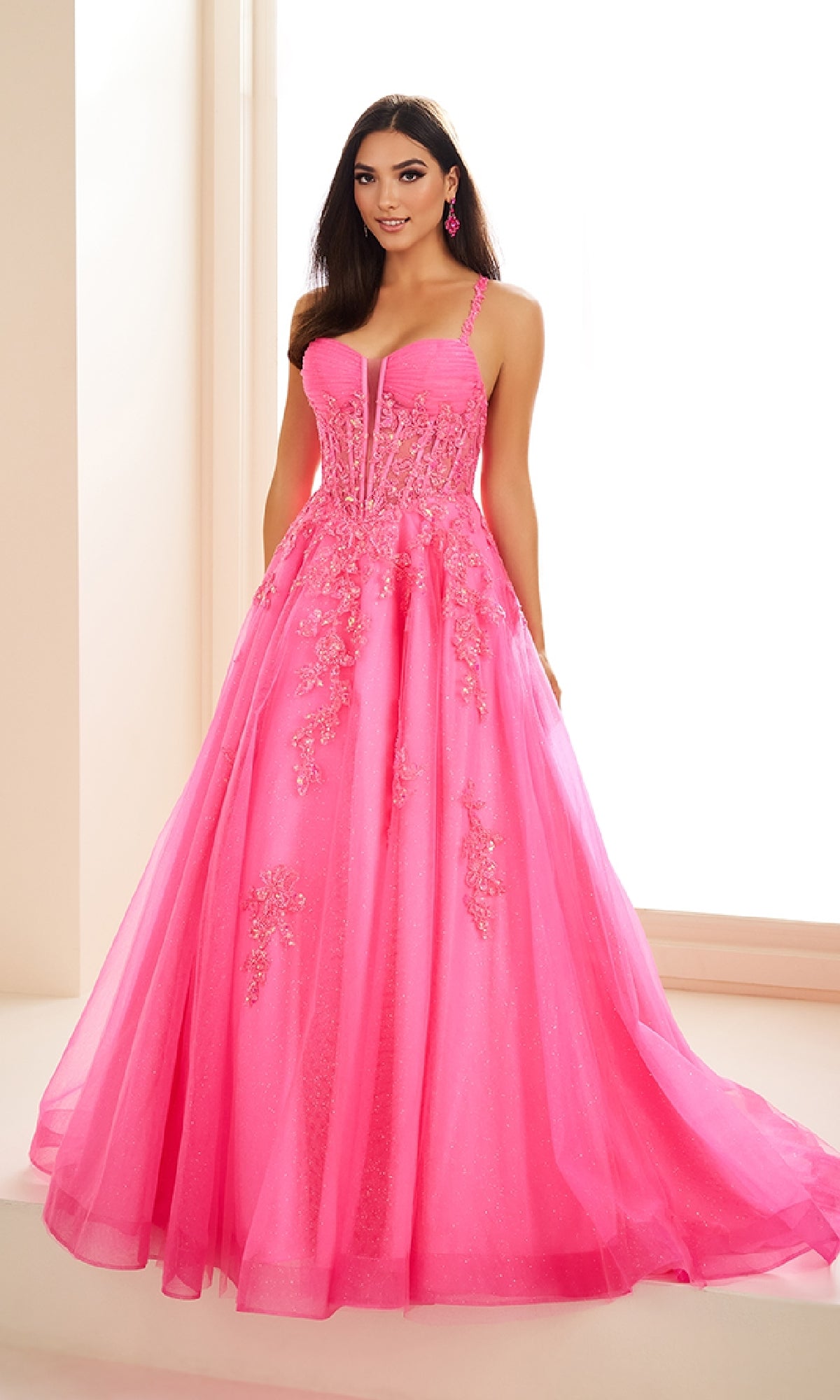 A woman stands indoors wearing the Ellie Wilde EW36202 long prom dress in bright pink with floral embroidery and a corset bodice, looking at the camera.