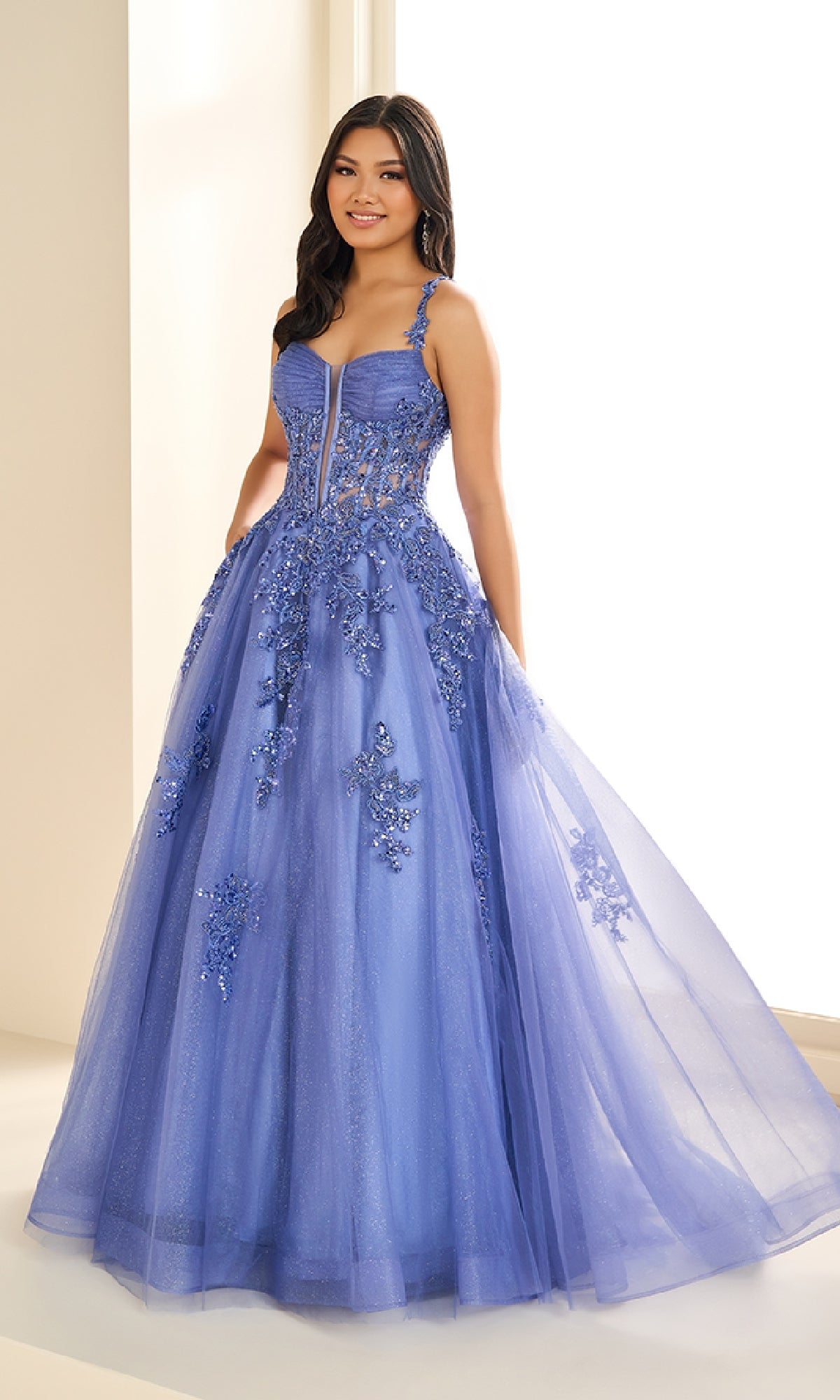 A young woman stands indoors wearing the Ellie Wilde EW36202 long prom dress, a floor-length sleeveless lavender gown featuring a corset bodice, delicate floral appliqués, and a sheer glitter tulle skirt.