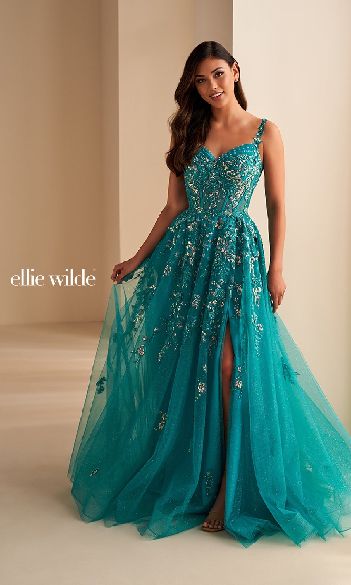 A woman wears the Ellie Wilde EW36204 long prom dress—a teal embellished ball gown with a corset bodice, thigh-high slit, and floral lace appliques—standing in a neutral room.